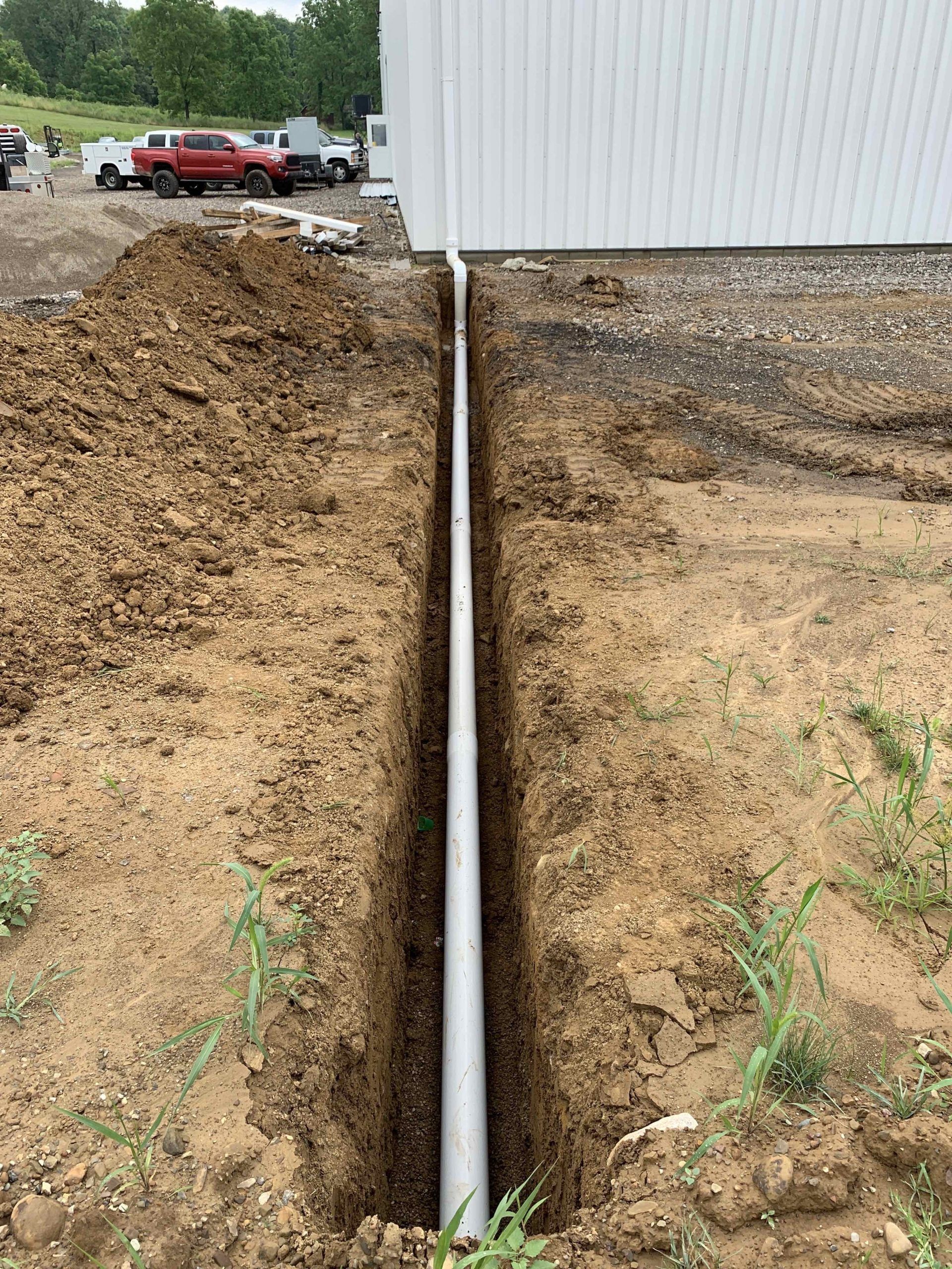 A narrow trench dug alongside a white building with a PVC pipe laid inside.