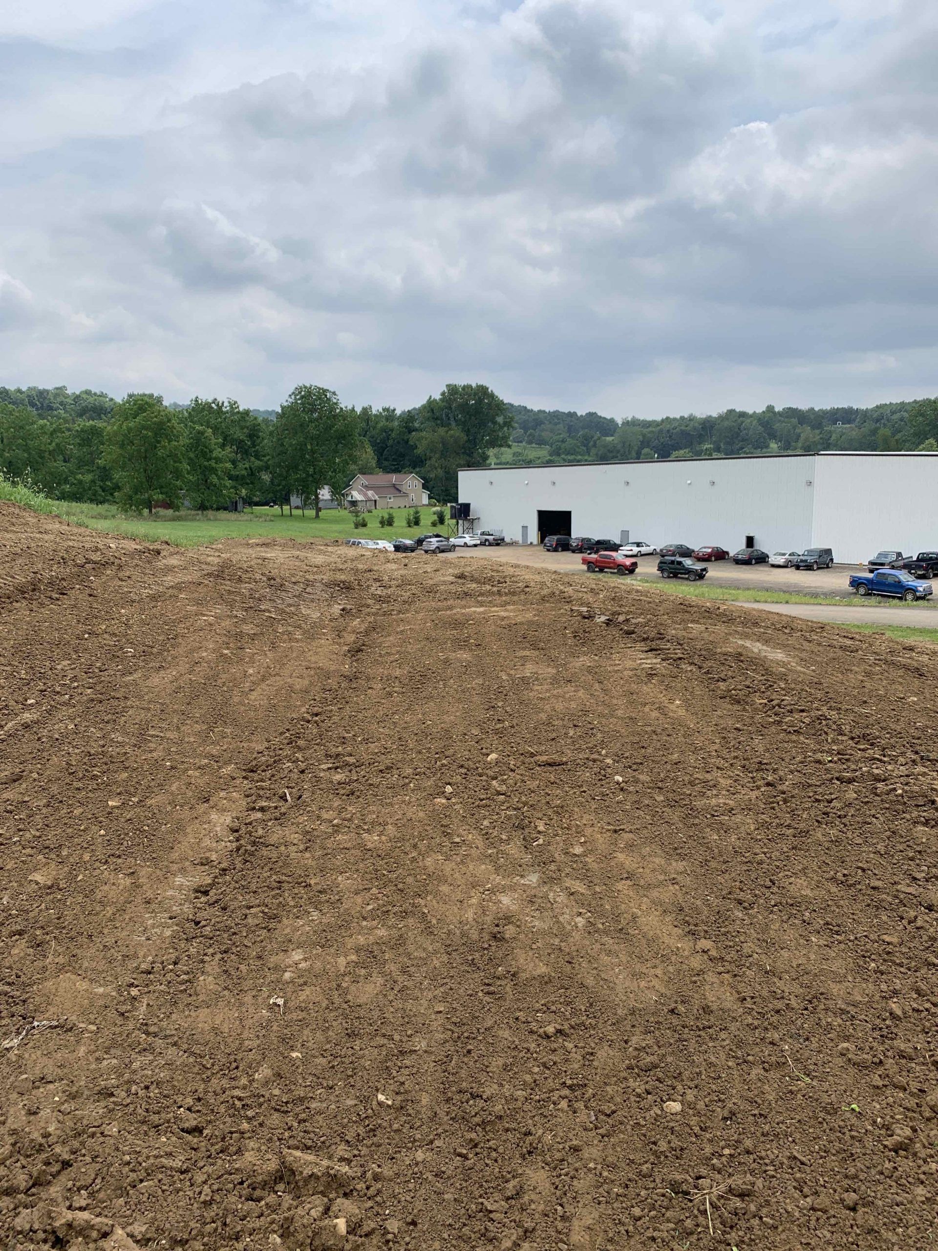 A pile of dirt in front of a white building with parked cars, a house, and trees under a cloudy sky.