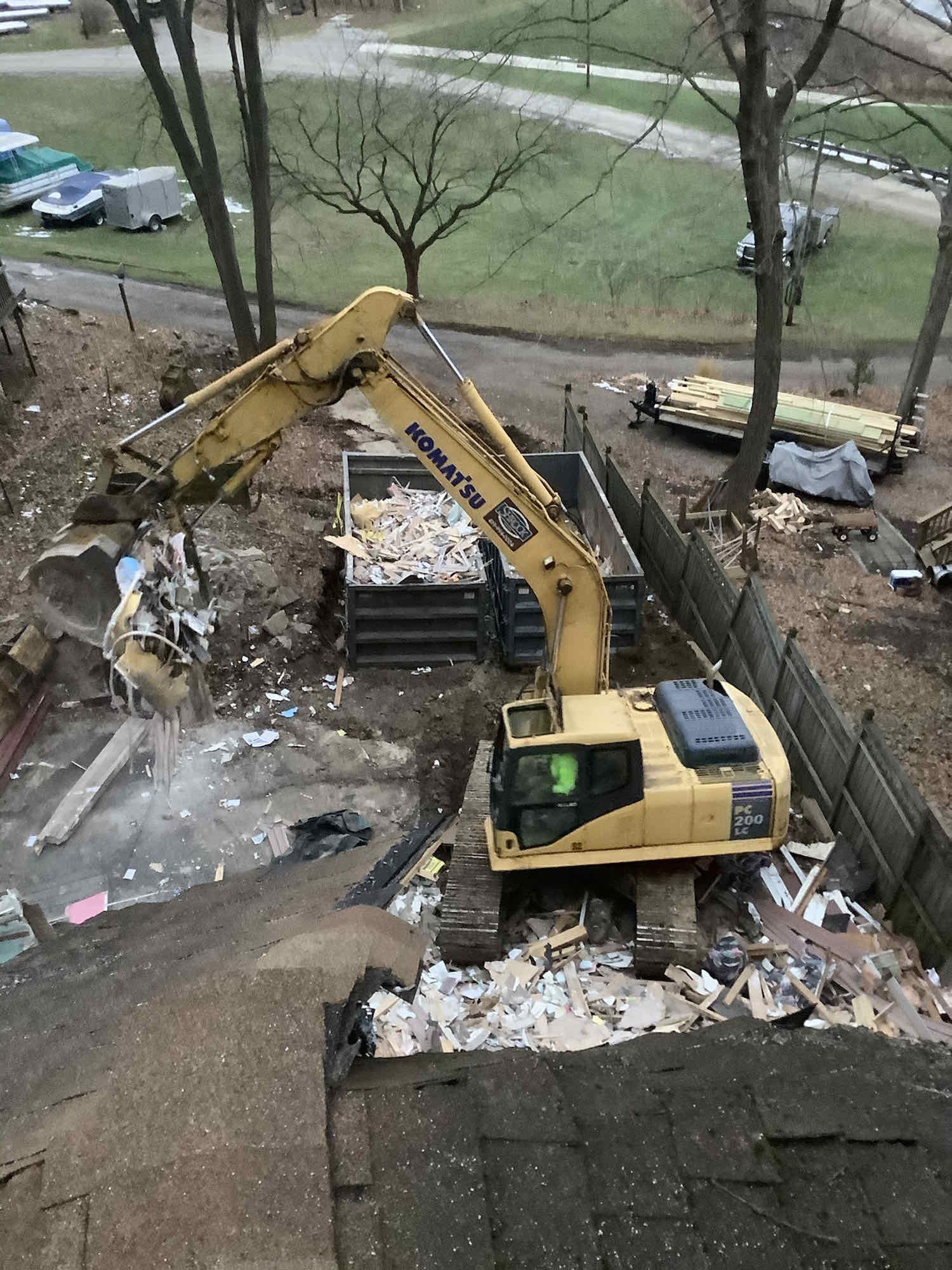 Yellow excavator demolishing a building, loading debris into a container. Overcast day.