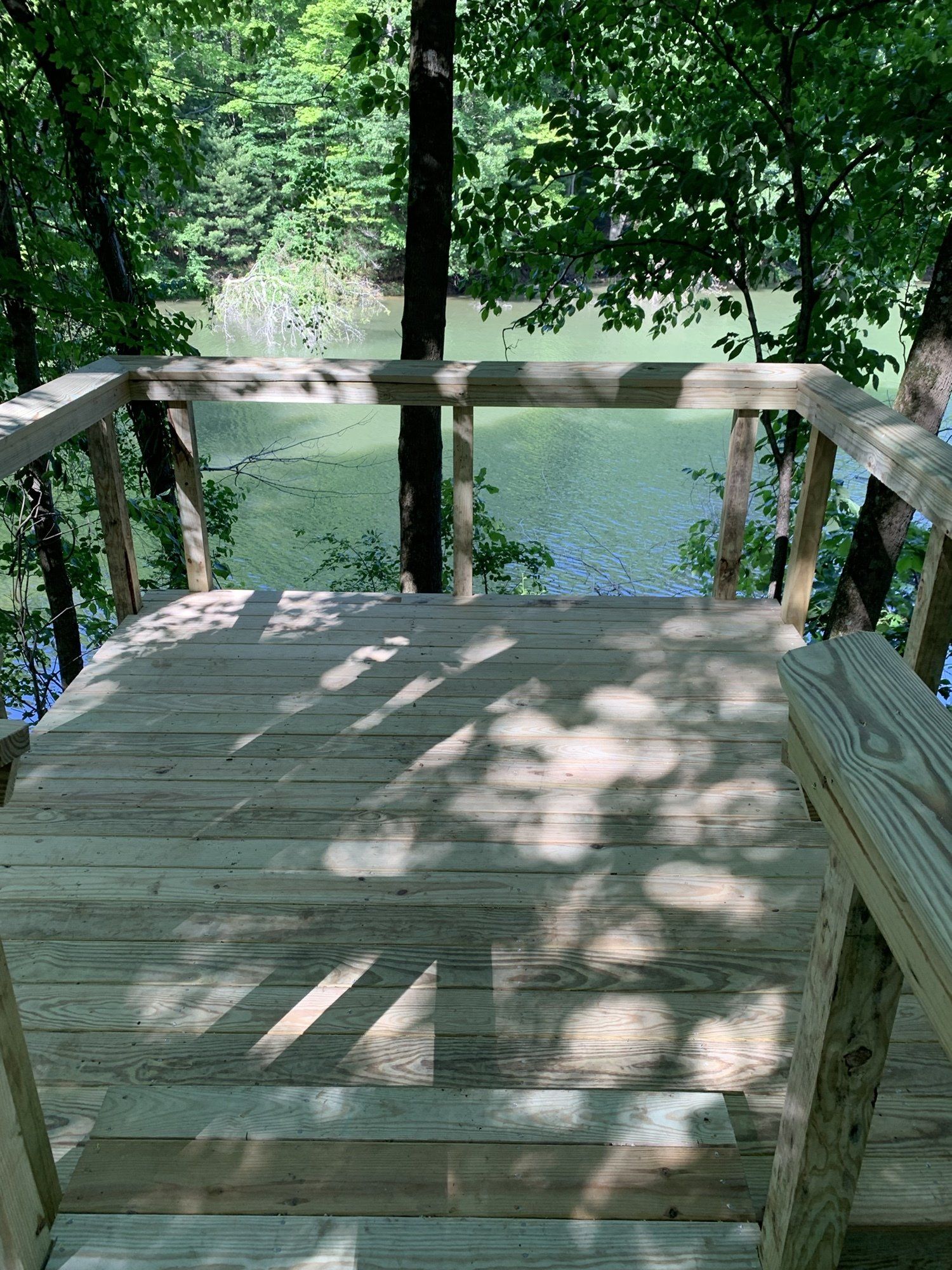 Wooden deck with railing overlooking a lake. Sunlight filters through trees, casting shadows on the deck.