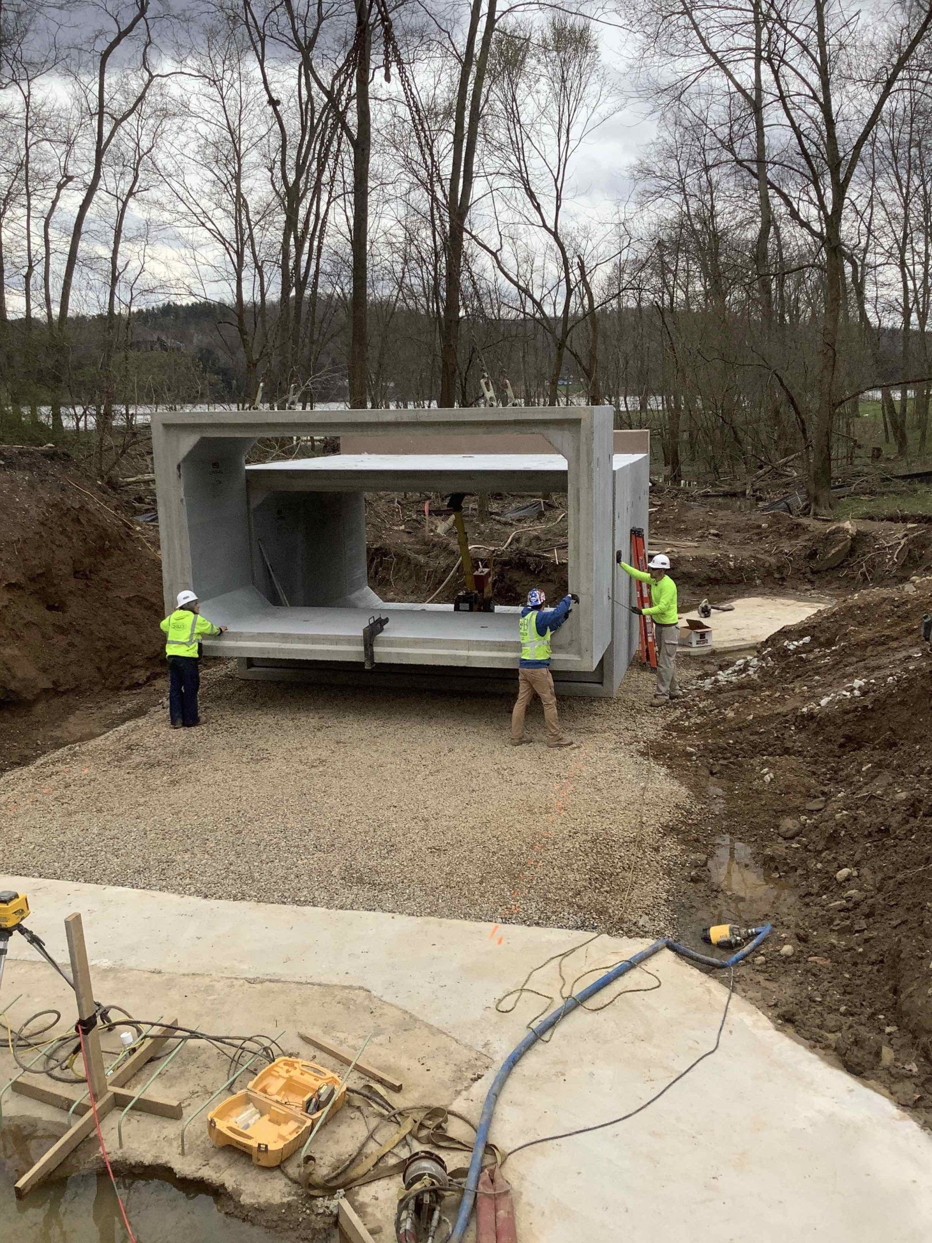 Construction workers positioning a large concrete box culvert on gravel. Setting is near trees and water.