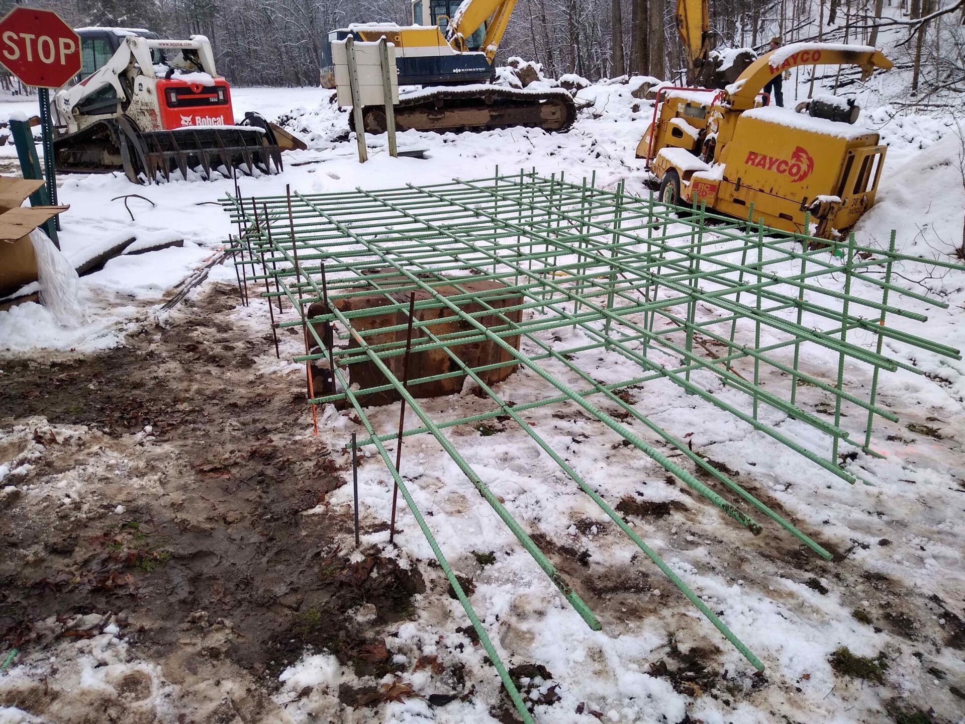 Construction site with rebar framework on snowy ground, heavy machinery in background.