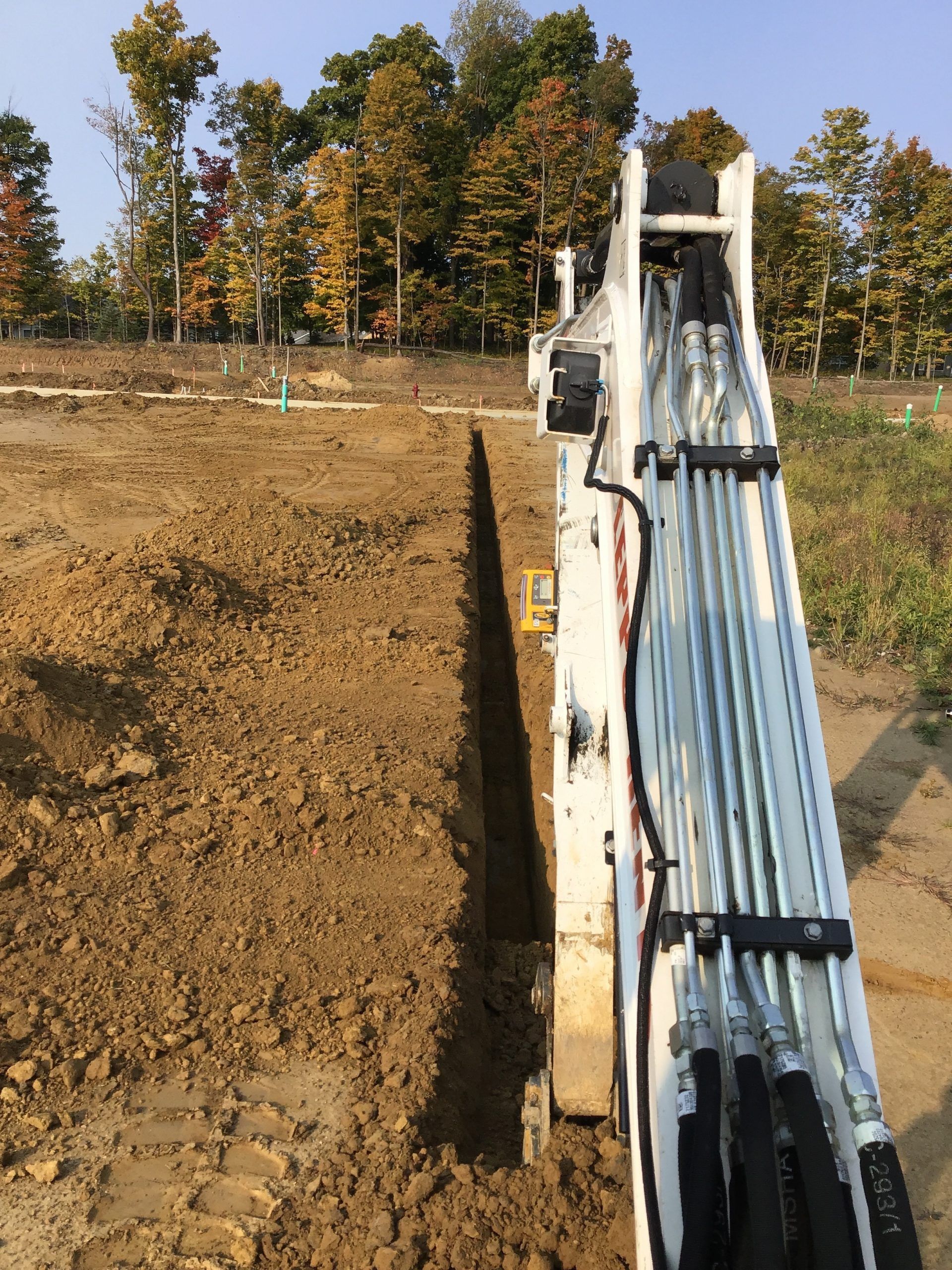 A white excavator digging a narrow trench in brown soil, with a forest in the background.