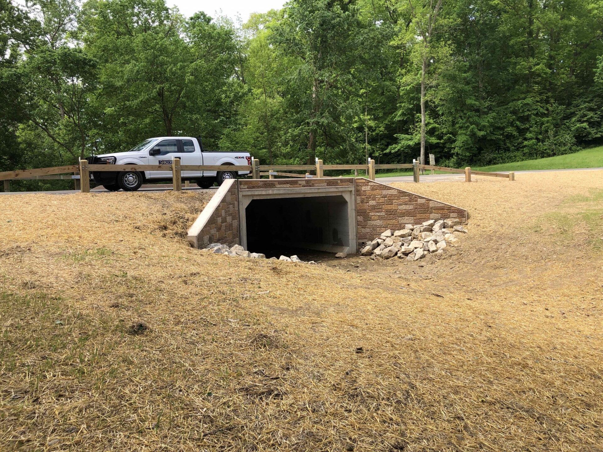 White pickup truck near a culvert tunnel under a road, surrounded by brown gravel, trees, and a wooden fence.