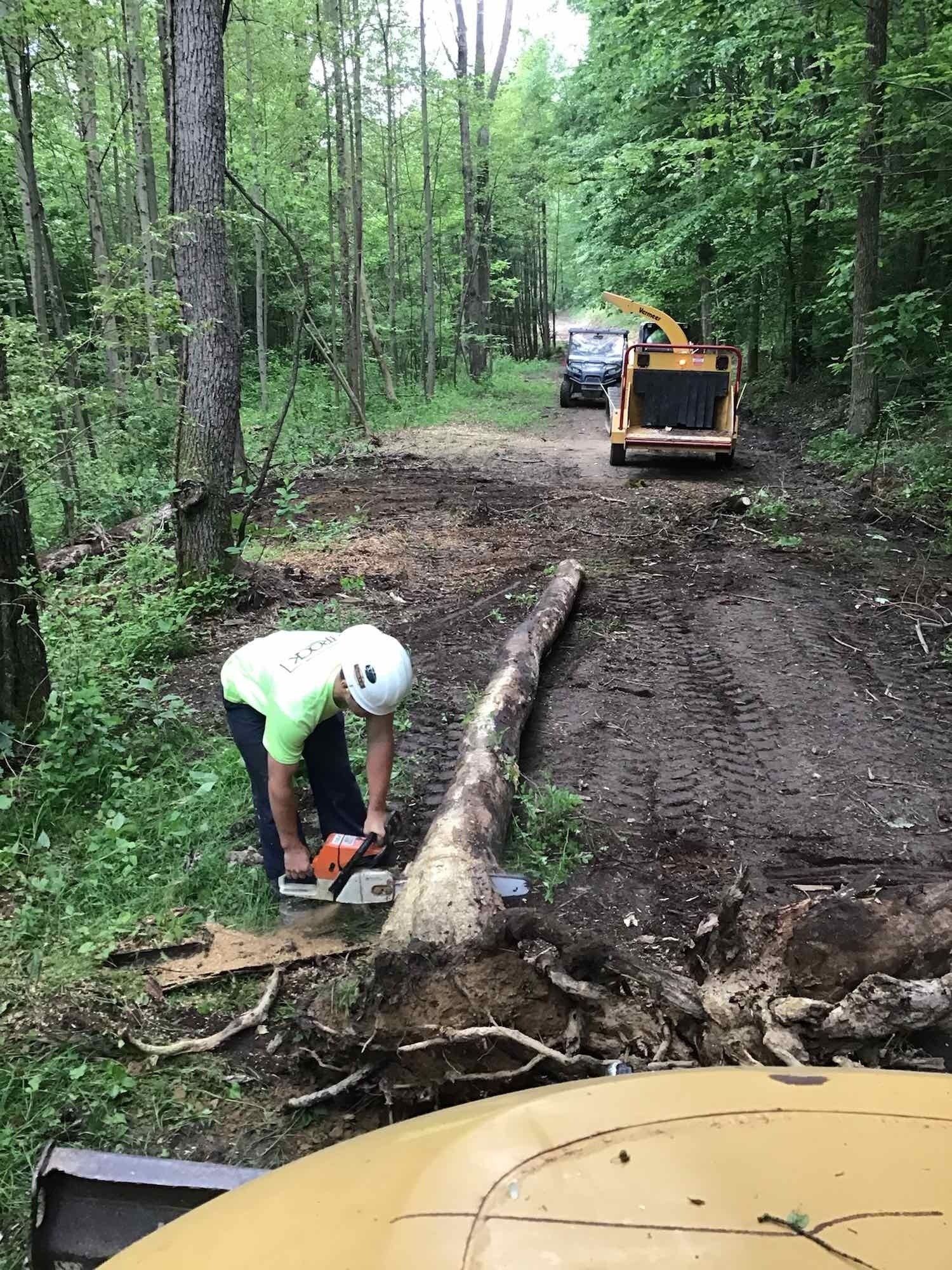 Man using a chainsaw to cut a log on a dirt road, with a wood chipper and truck in the background.