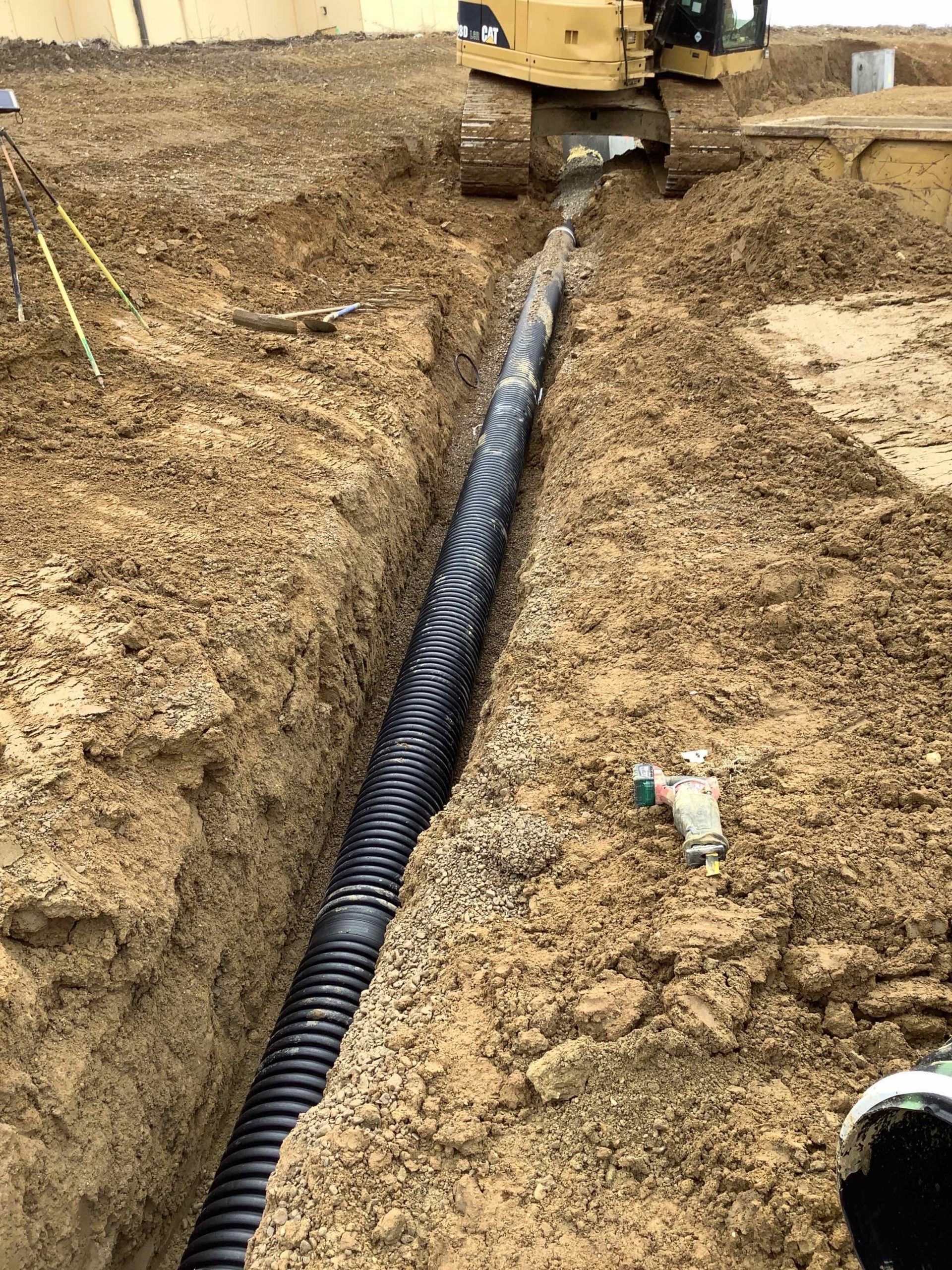 Black corrugated pipe in a trench with a yellow excavator at a construction site.