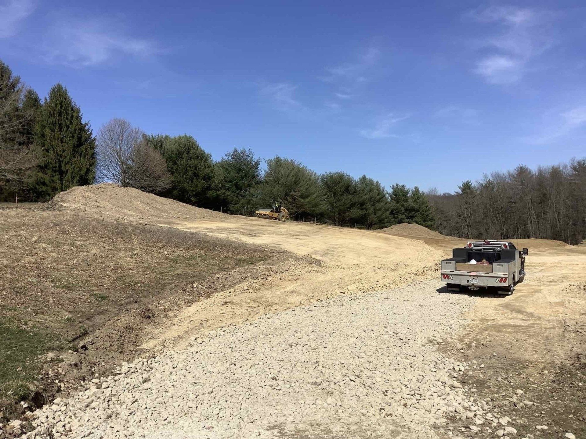Construction site with gravel road, dirt piles, trailer, and trees under a blue sky.