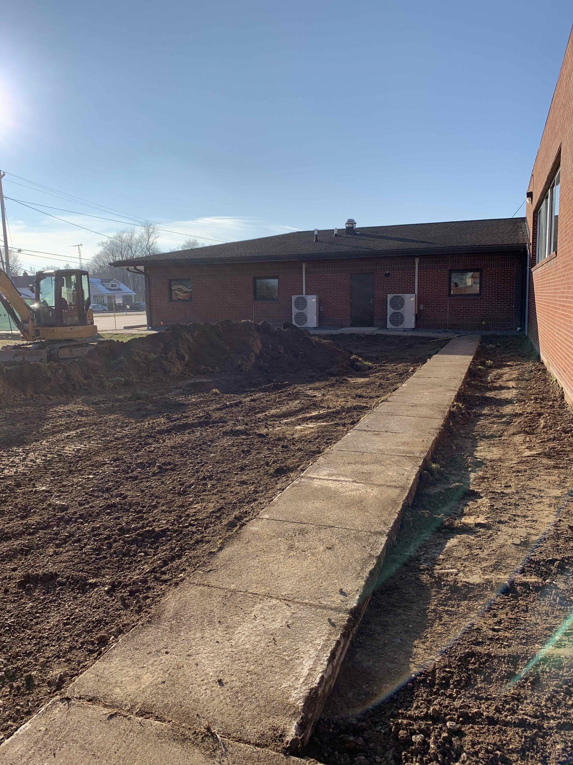 Exterior view of a building with a concrete walkway. Excavation work is visible in front. Clear, sunny day.