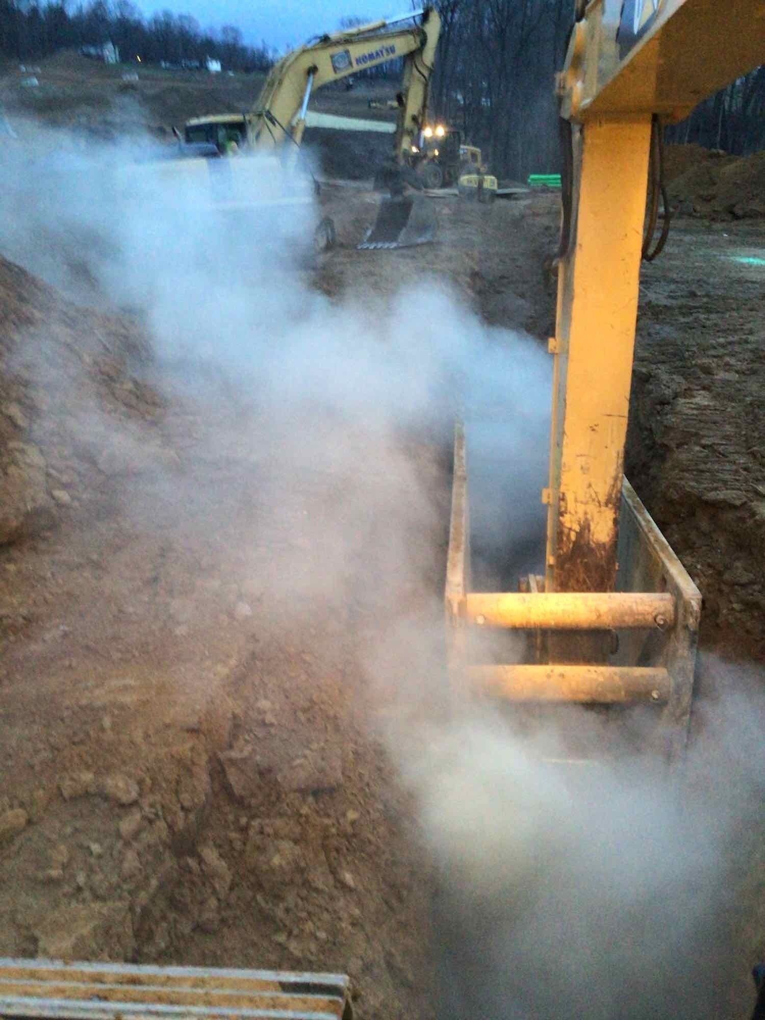 Yellow excavator digging a trench, releasing steam or dust. Another excavator in the background. Construction site.