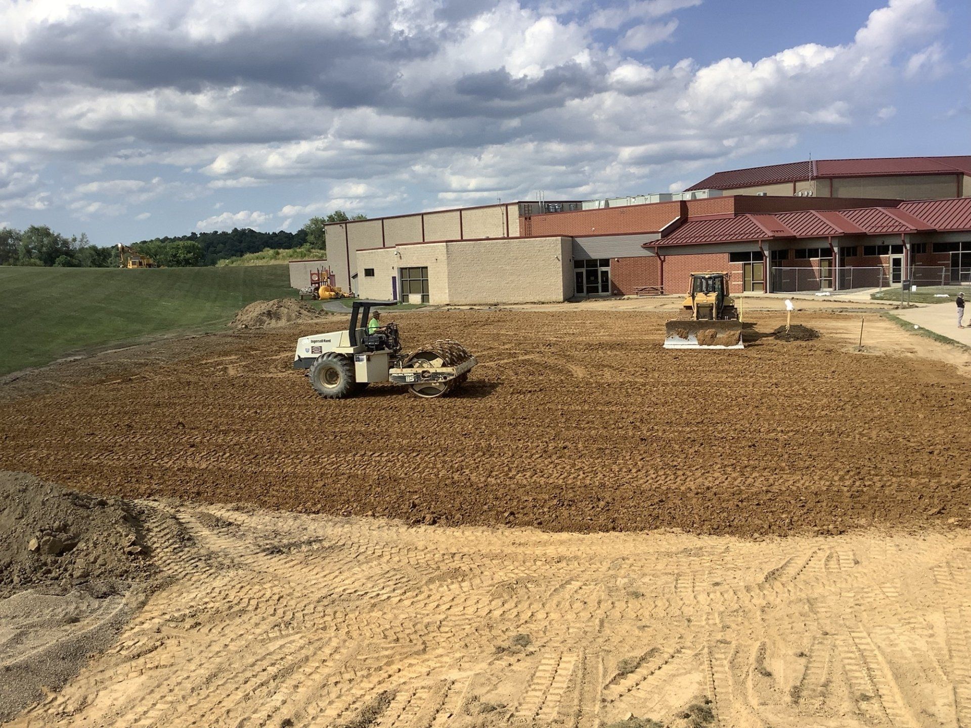 Construction site. A roller and bulldozer compact brown soil in front of a large building. Cloudy sky.
