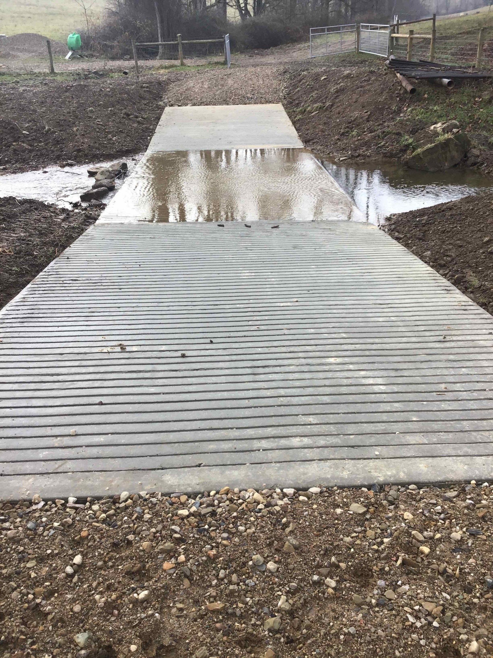 Gray concrete walkway over shallow water, between gravel banks, leading towards a green hillside.