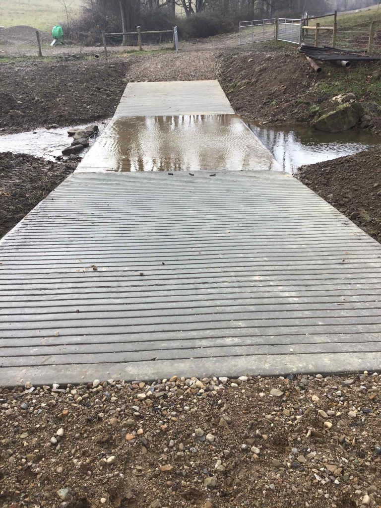 A wooden-style bridge partially covered in water, surrounded by dirt and vegetation.
