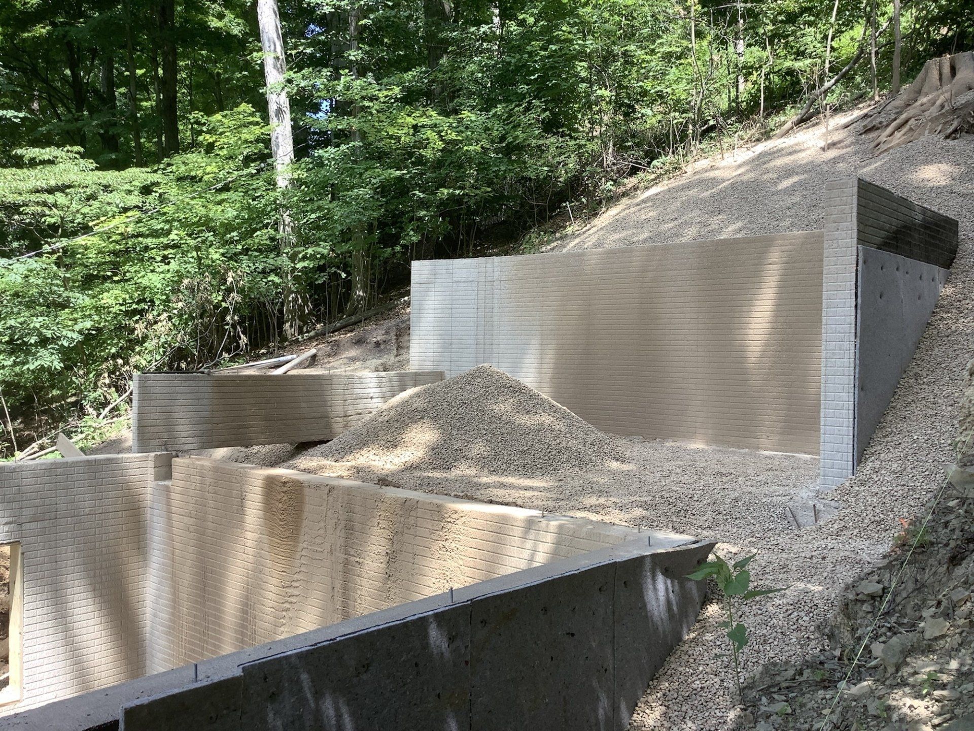 Concrete retaining walls supporting a hillside, filled with gravel. Green foliage in the background.