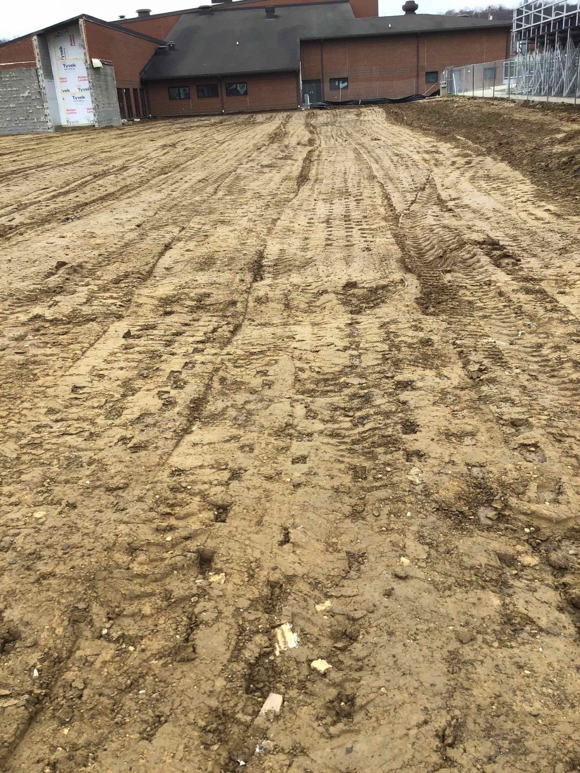 Muddy, tilled earth. Buildings are in the background. Tracks from equipment are visible.