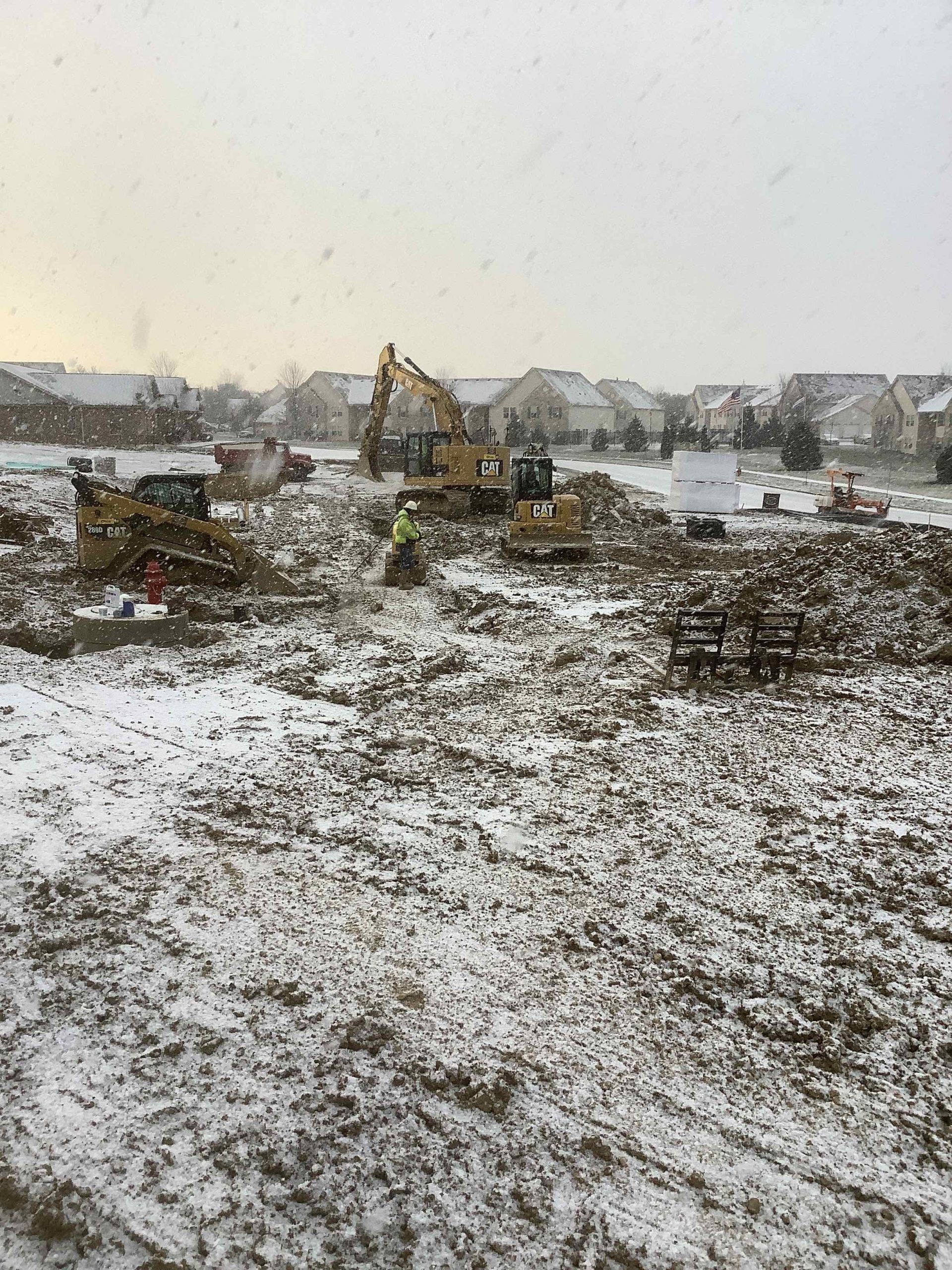 Construction site covered in snow, with excavators and a worker present. Residential buildings in background.