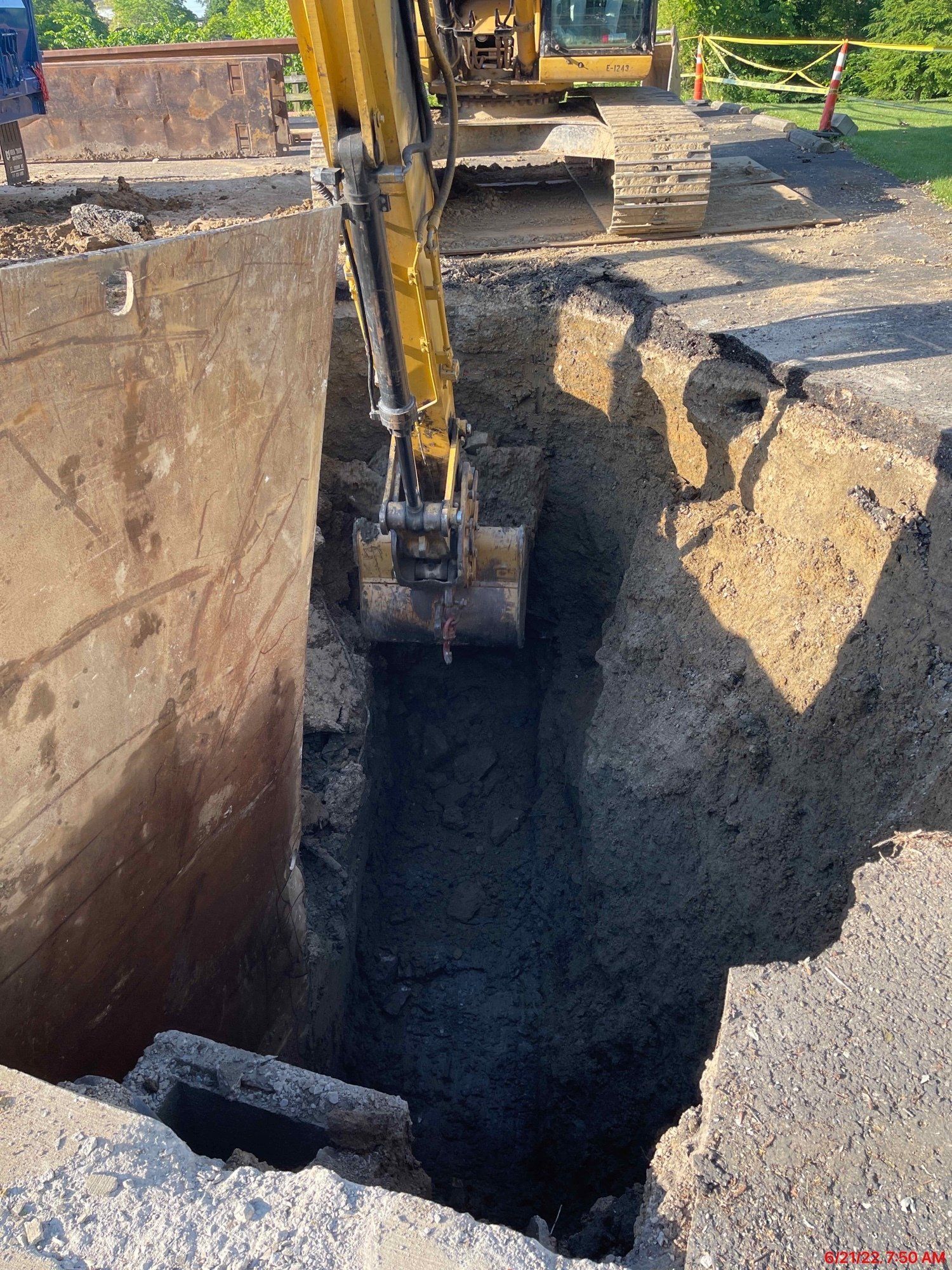 An excavator digs a deep trench in asphalt. The edge of a concrete structure is visible on the left.