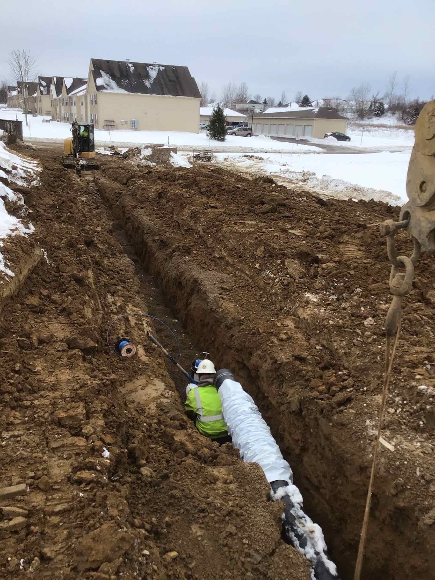 Construction worker in a trench with pipe, snowy ground, and houses in the background.