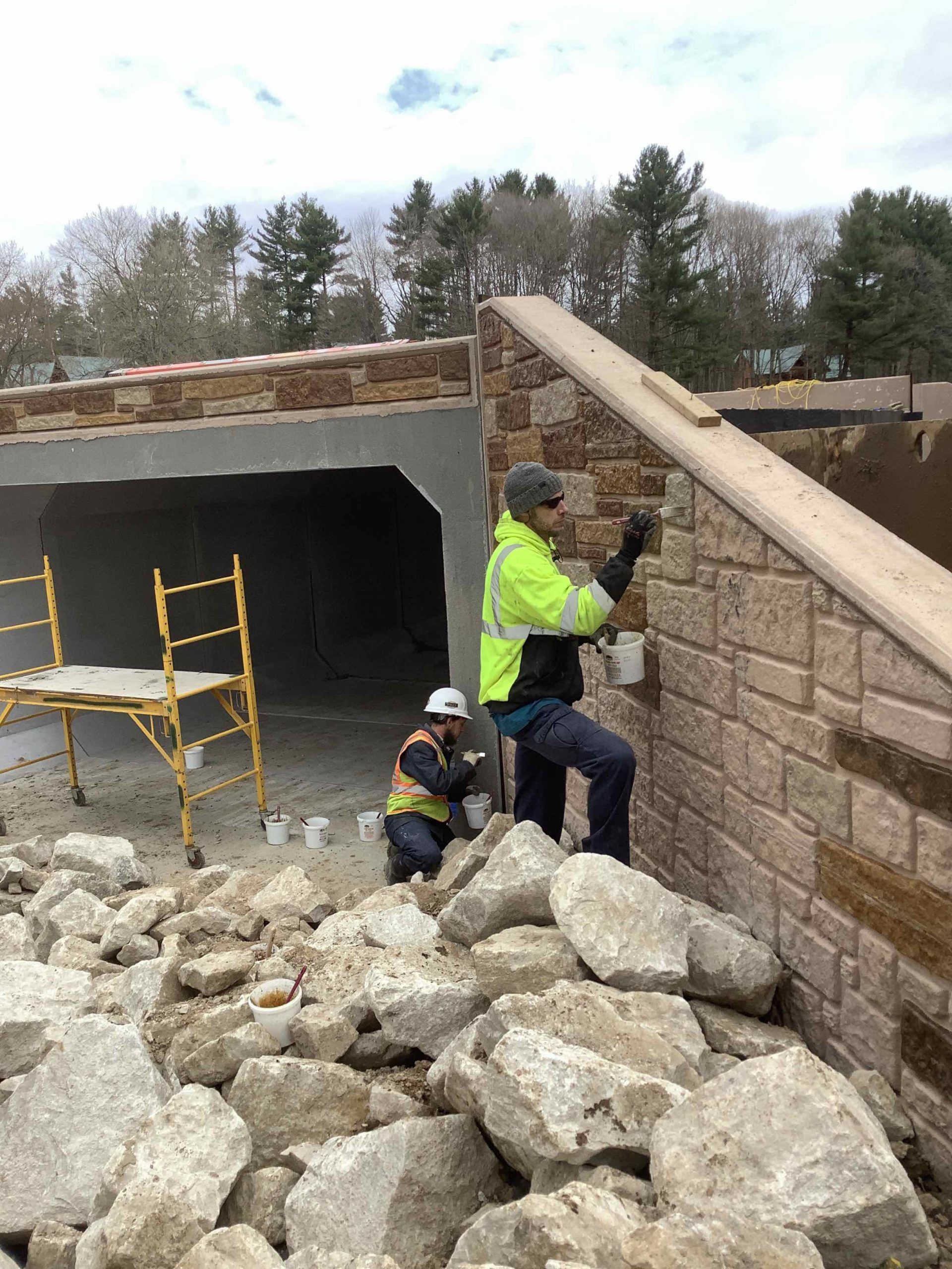 Two construction workers applying stone to a wall; one standing, one kneeling, debris in foreground.