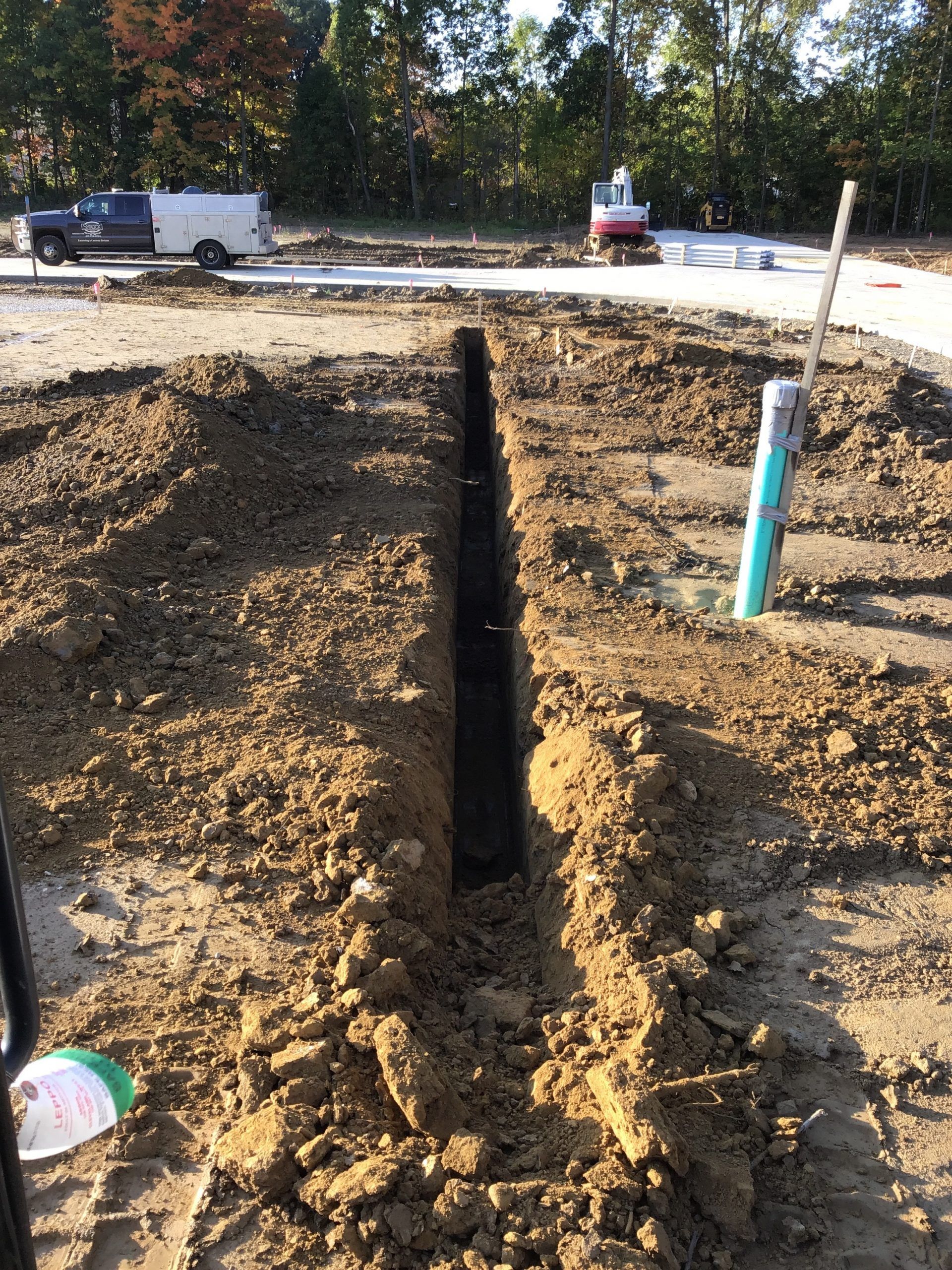 Trench dug in dirt, with a black pipe inside. Blue pipe and vehicles in the background.