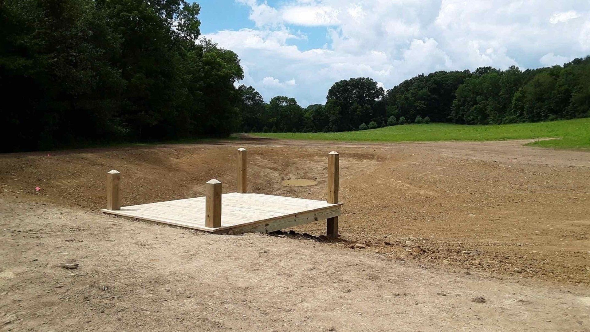 Wooden dock on dry, cracked earth; trees and green field in background under a blue sky.