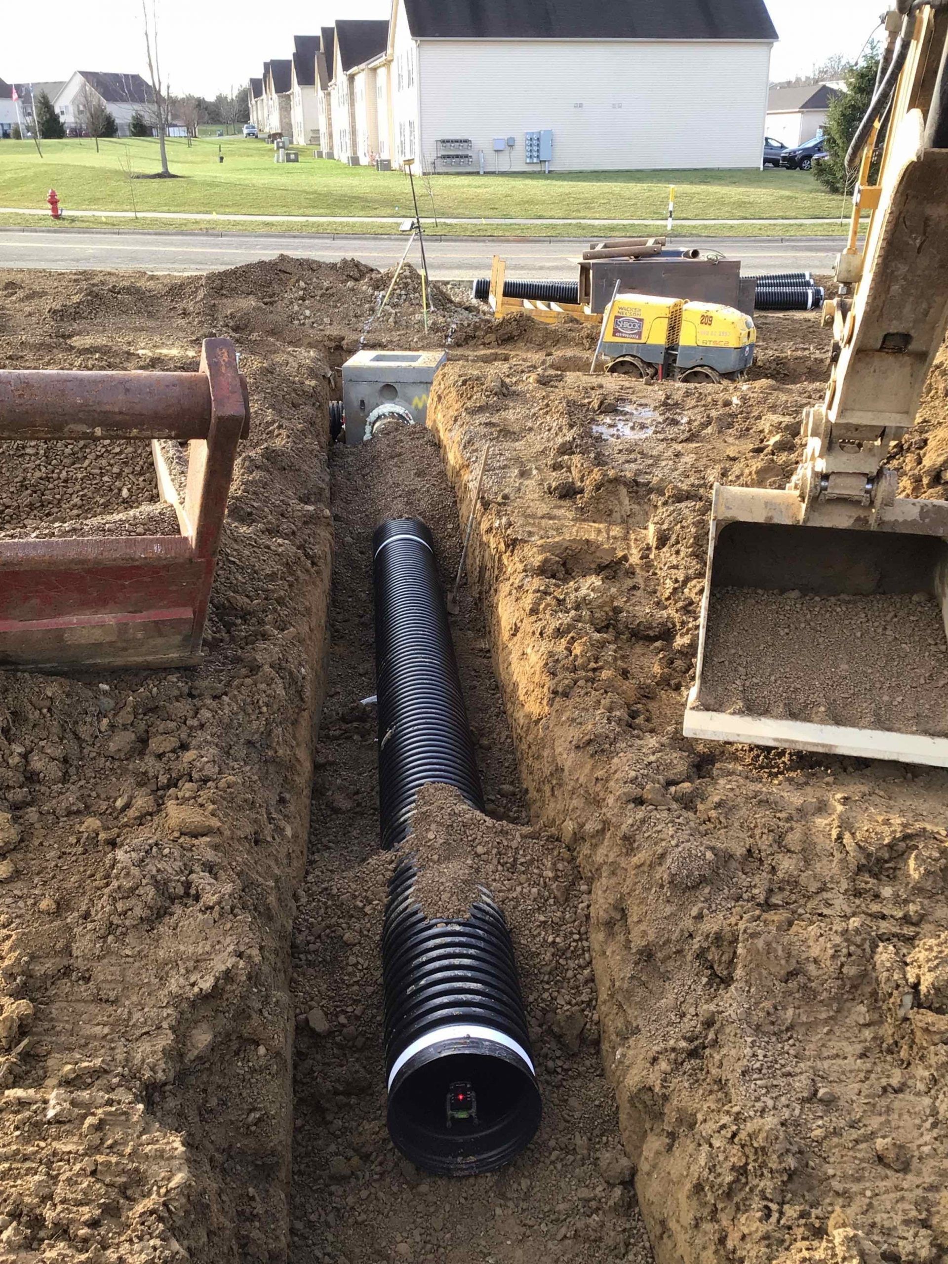 Trench dug for a large black pipe, with an excavator on the right, near a street and houses.