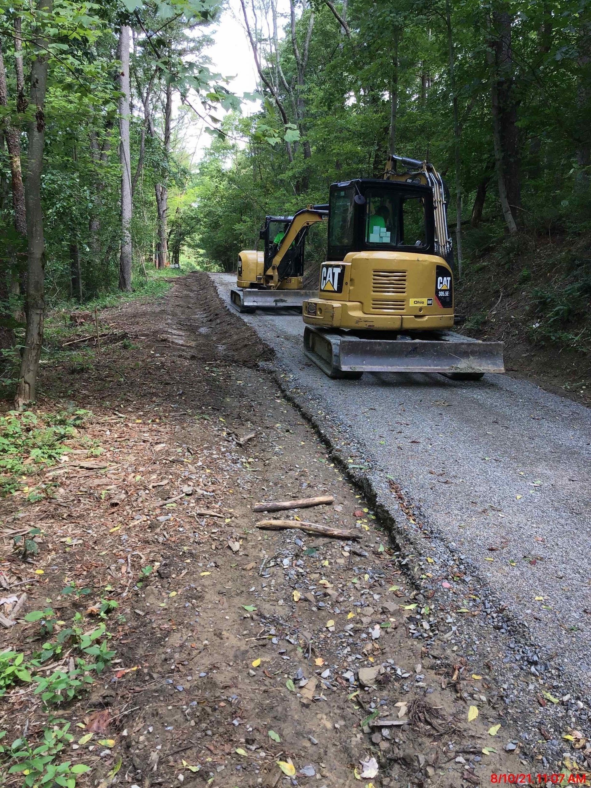 Two yellow Caterpillar mini-excavators on a gravel path in a forest. One is in the foreground.