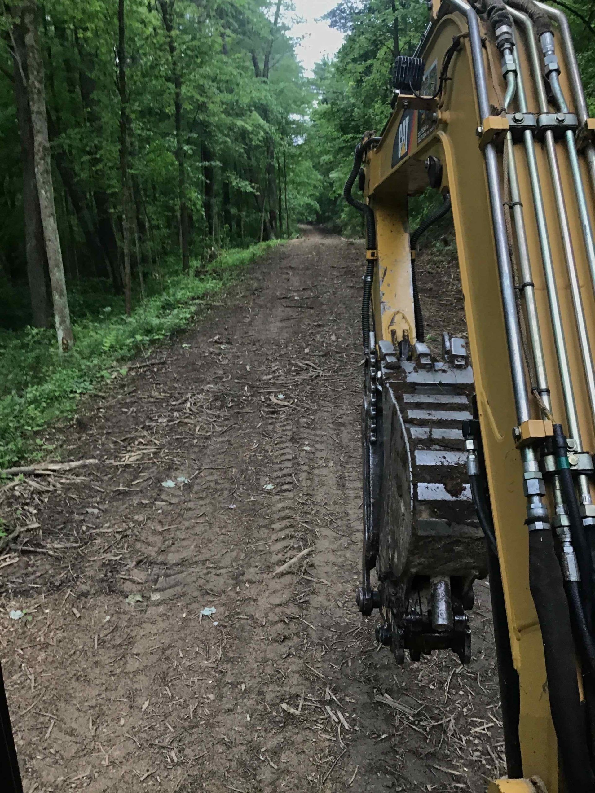 Excavator on a newly graded dirt path through a wooded area.