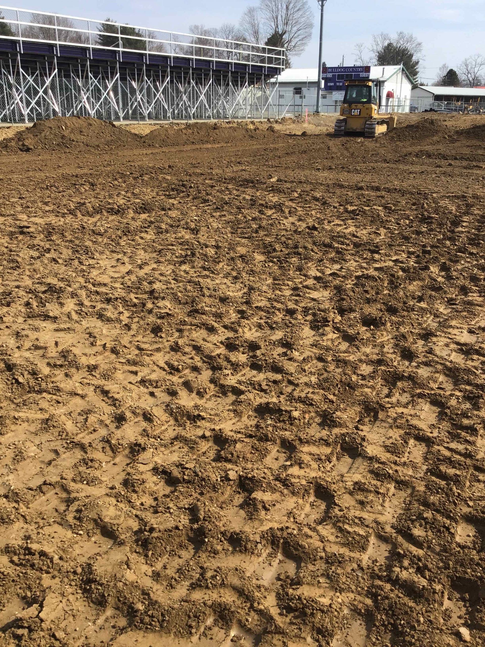 Muddy ground with a small bulldozer; bleachers and buildings in the background.