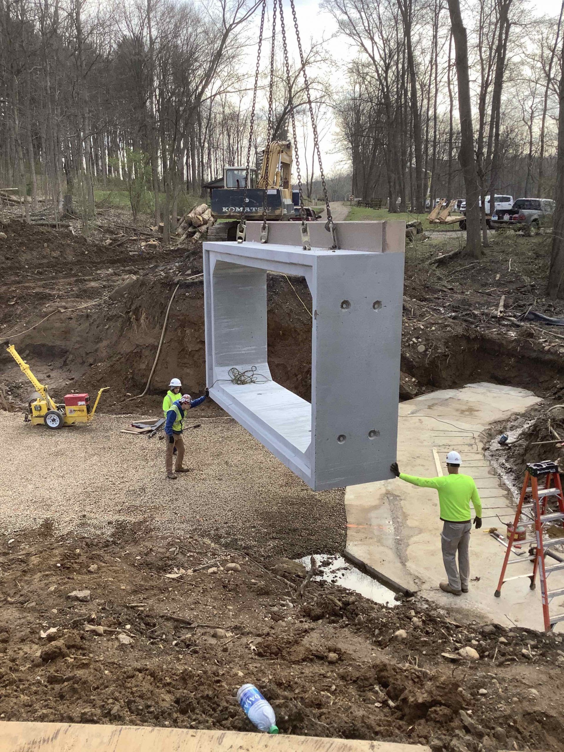 Large concrete culvert being lifted by a crane during construction. Workers in safety vests guide the structure.