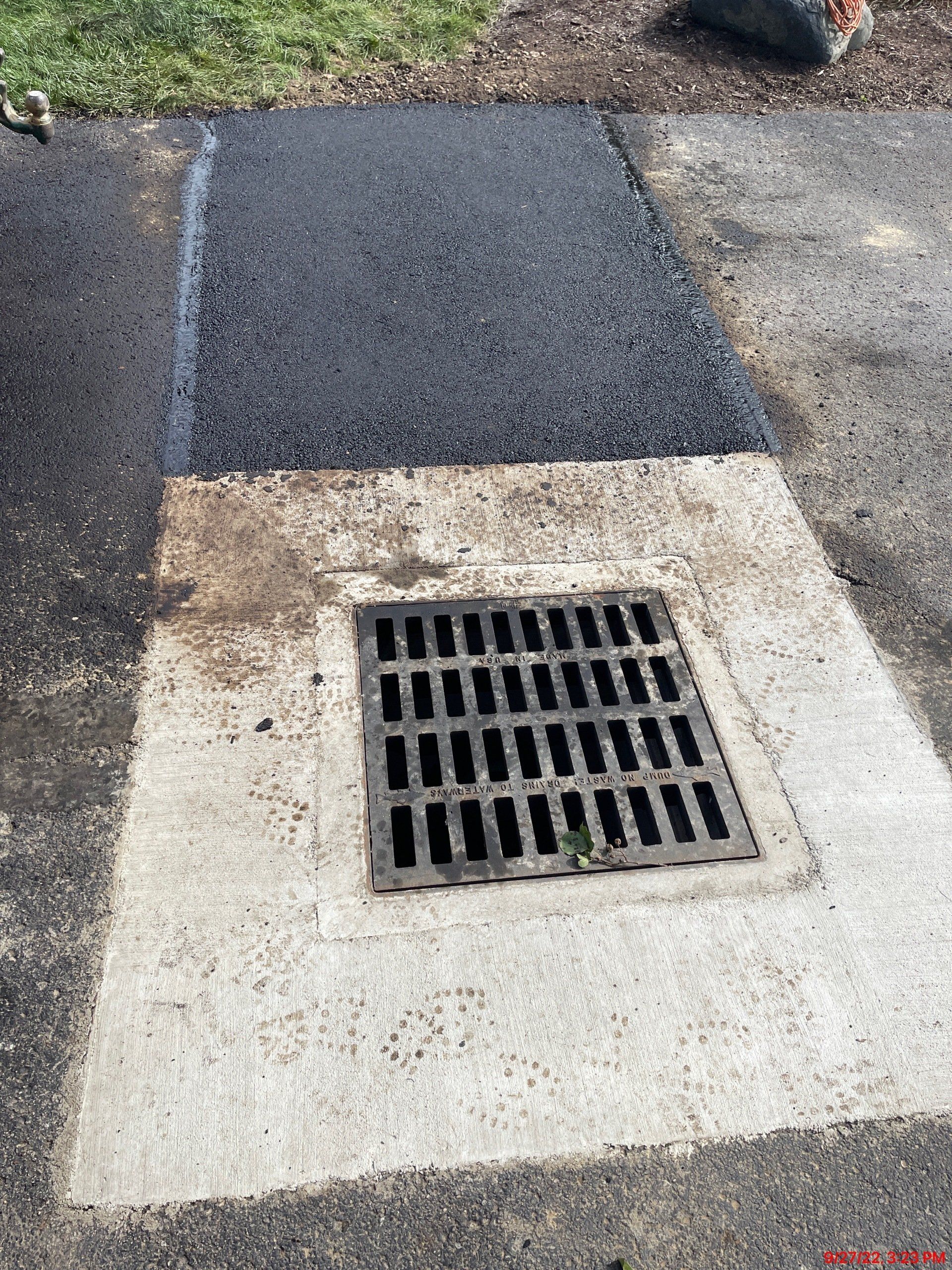 Asphalt patch next to a concrete drain, on an asphalt surface. Green grass visible.