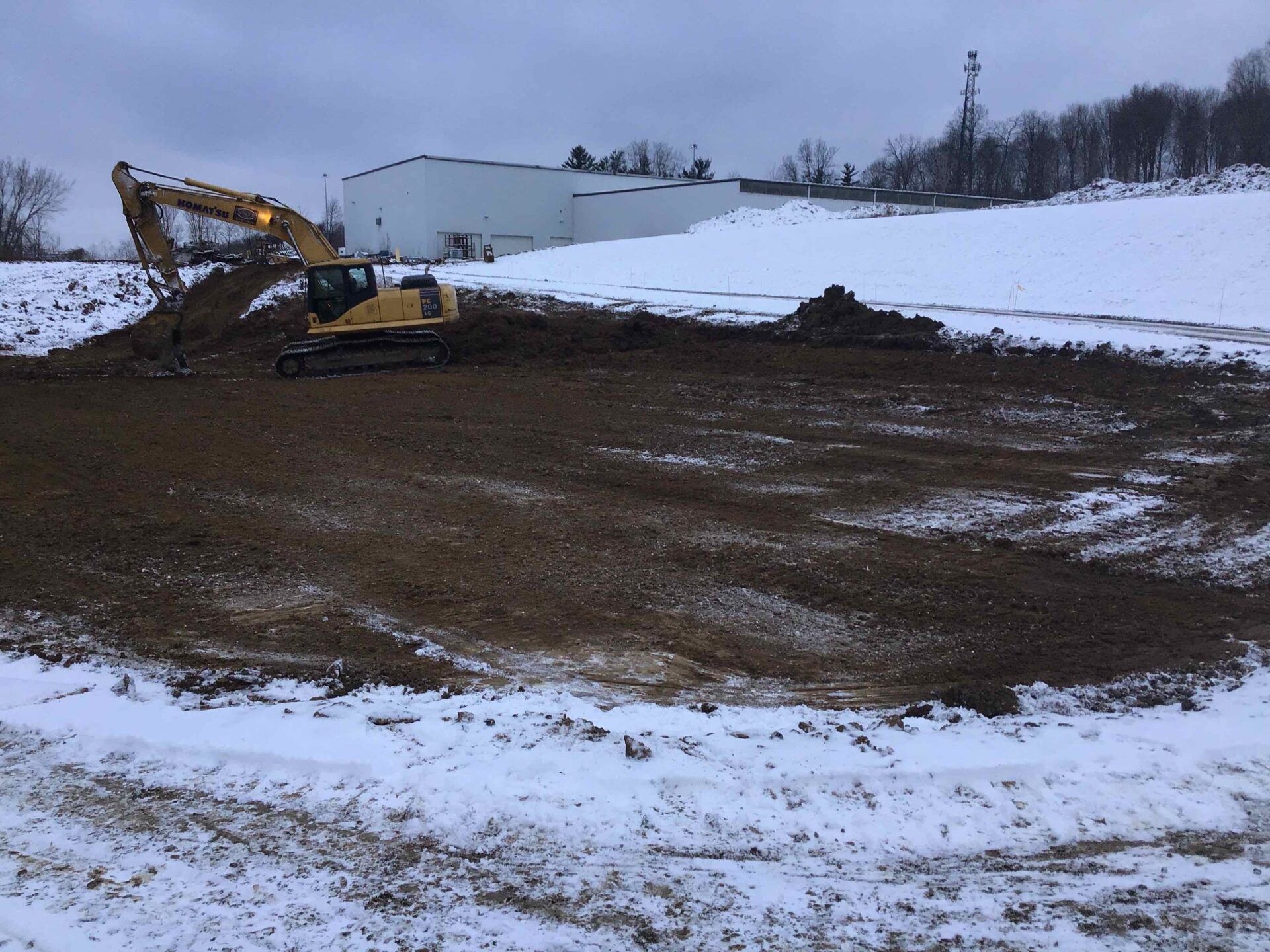 Excavator working on dirt pile in a snow-covered field. Large white building in the background.