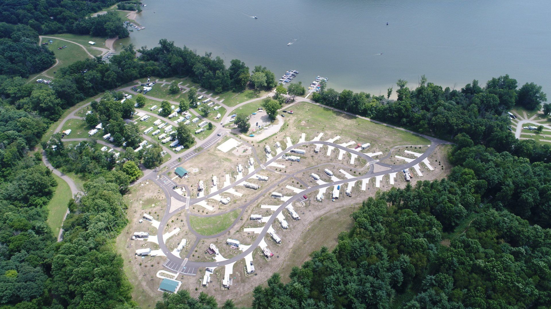 Aerial view of a campsite near a lake with numerous RVs and tents amidst trees.