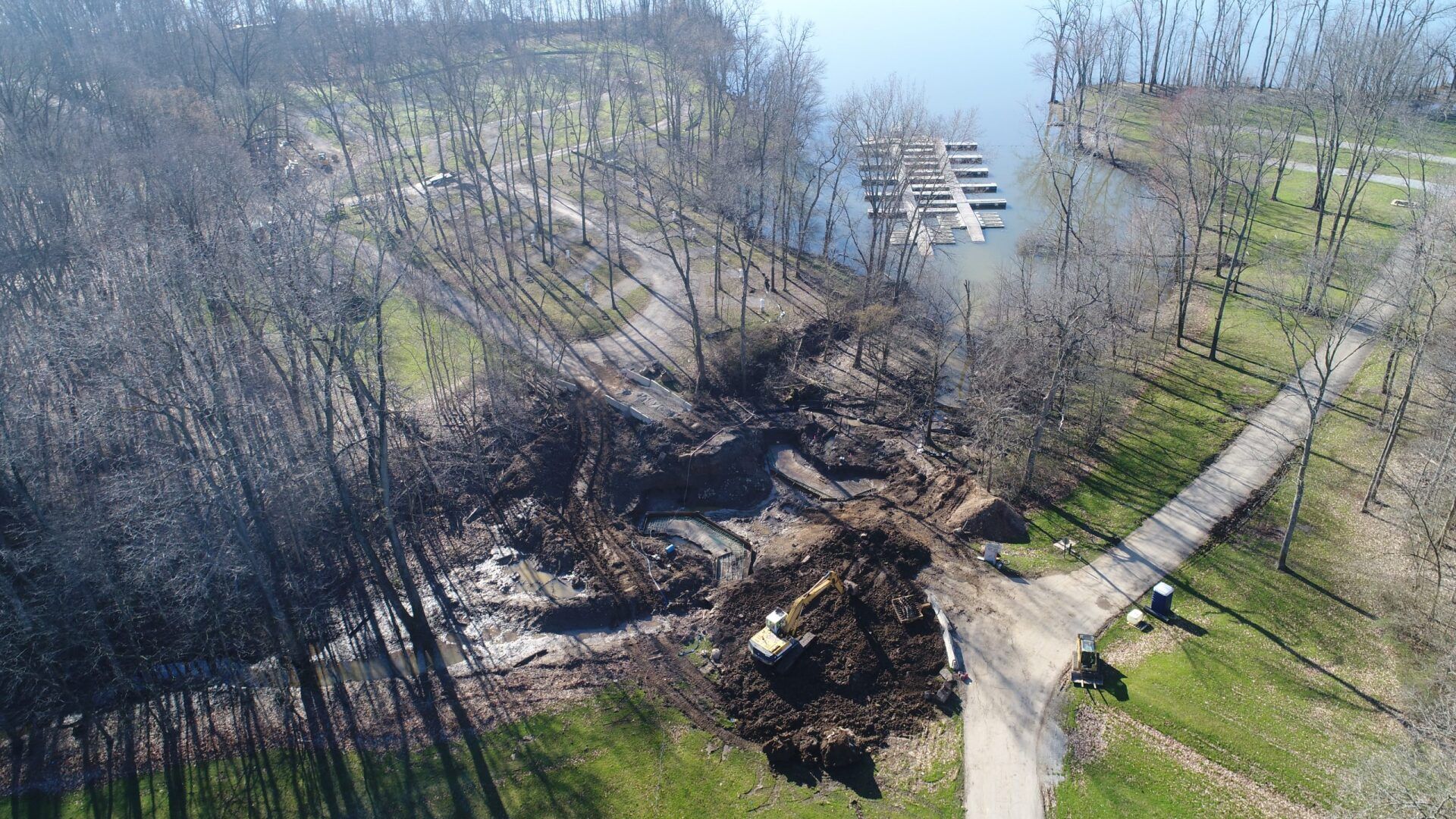 Aerial view: construction site with excavator, clearing a path near a marina and leafless trees.
