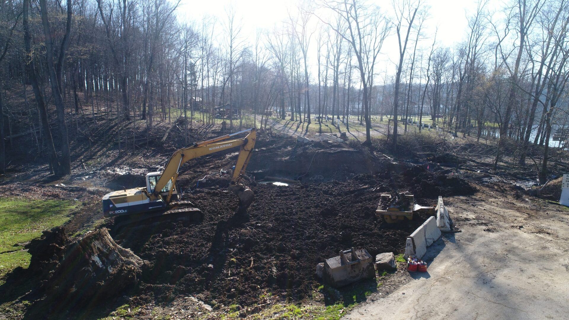 An excavator digs in a muddy area near a treeline. Bright sunlight.