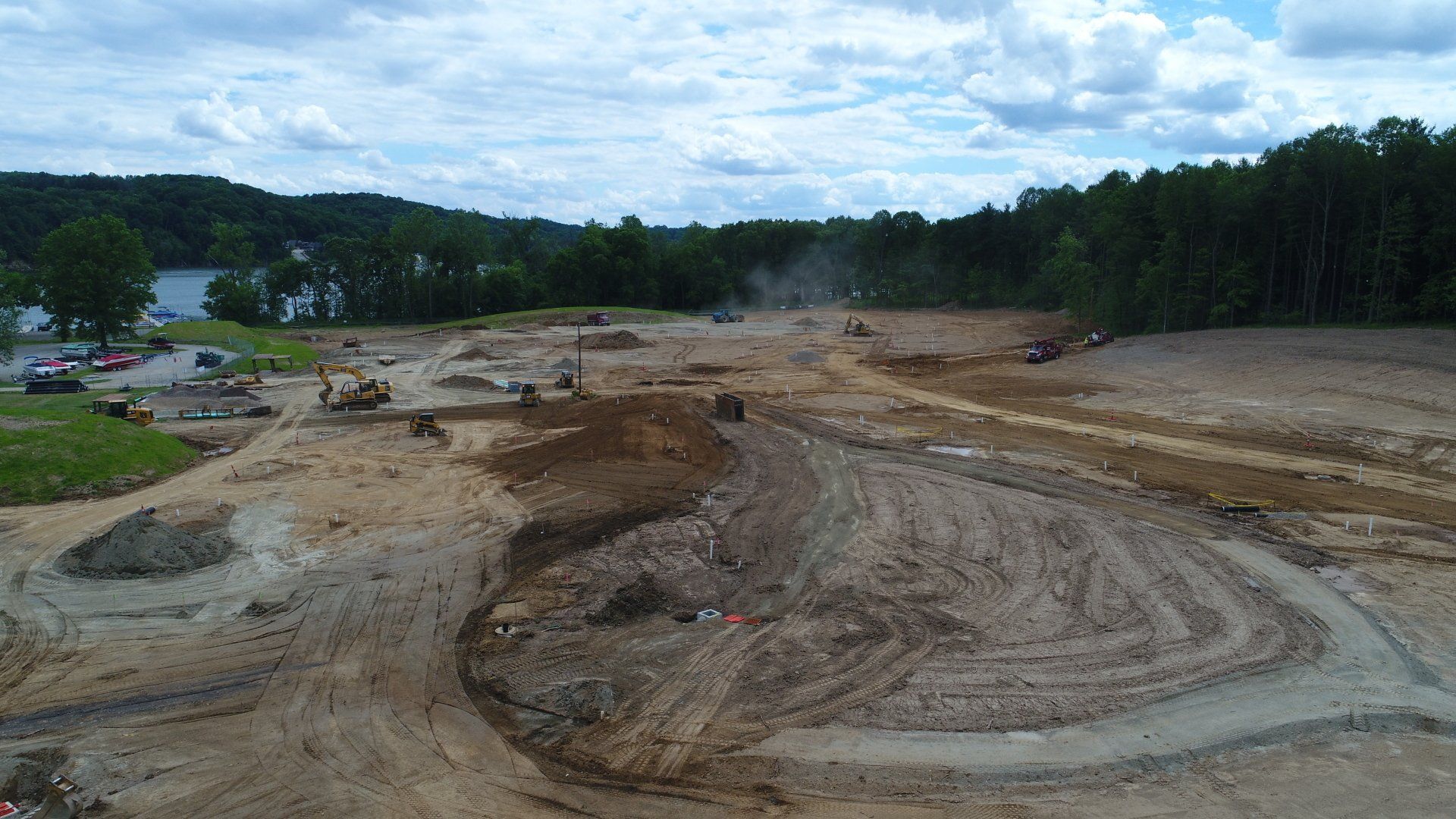 Construction site with heavy machinery, dirt piles, and a treeline in the background under a cloudy sky.