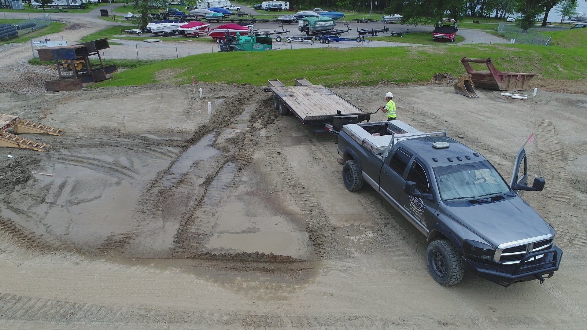 Dark pickup truck towing a trailer on a muddy construction site. A person in safety gear stands on the trailer.