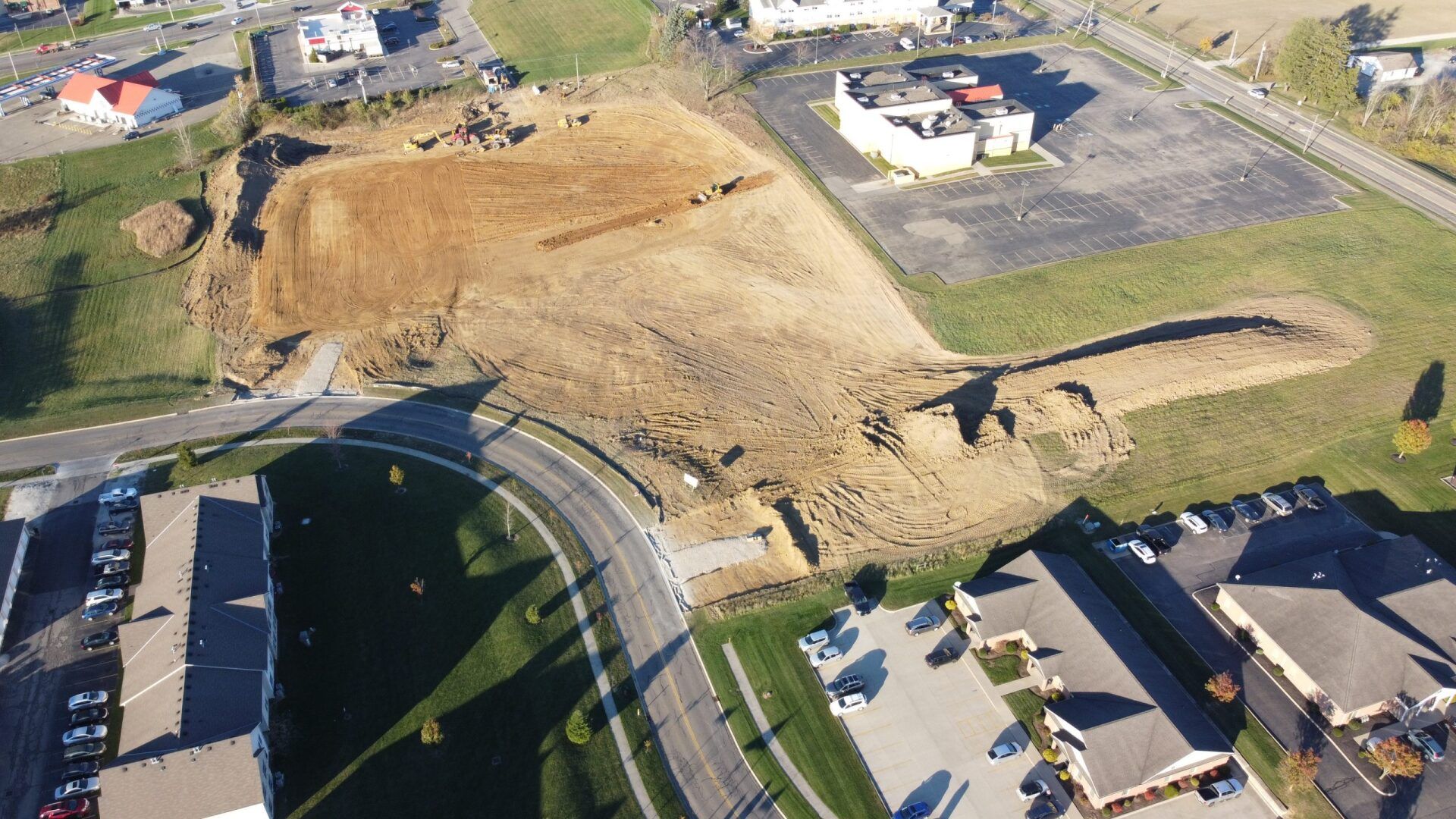 Aerial view of construction site with exposed dirt and surrounding buildings. Road curves in foreground.