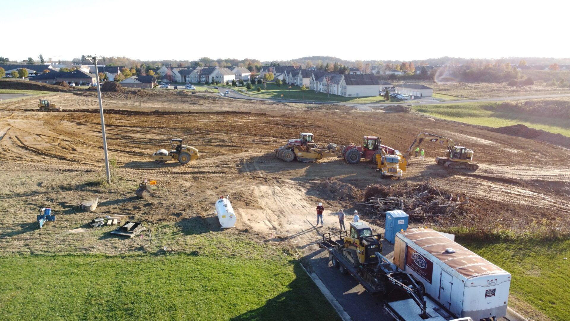 Construction site with heavy machinery, power pole, and mobile offices. Field in foreground, houses in background.