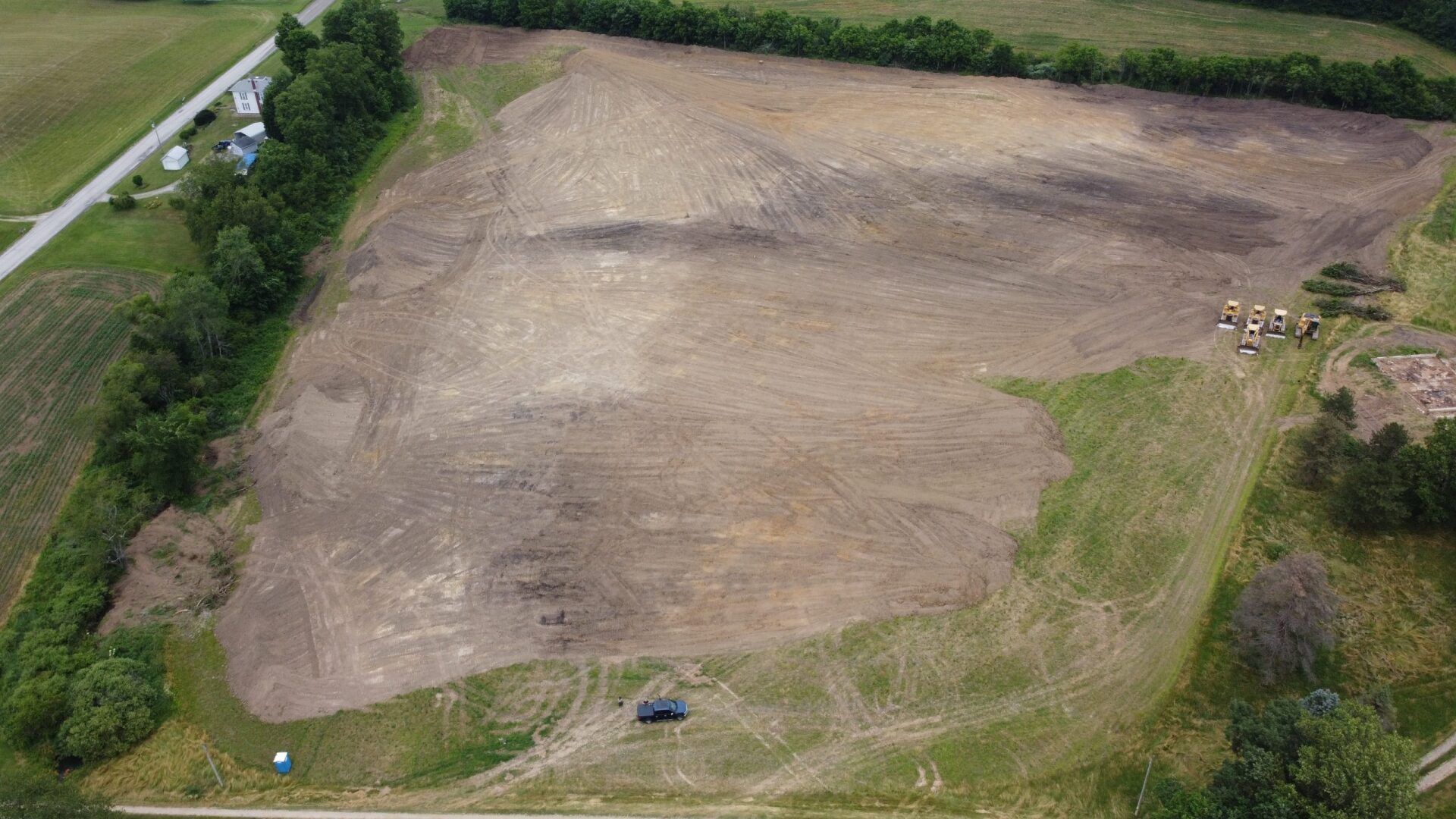 Cleared field of brown dirt, bordered by green trees. Road and small structures visible.