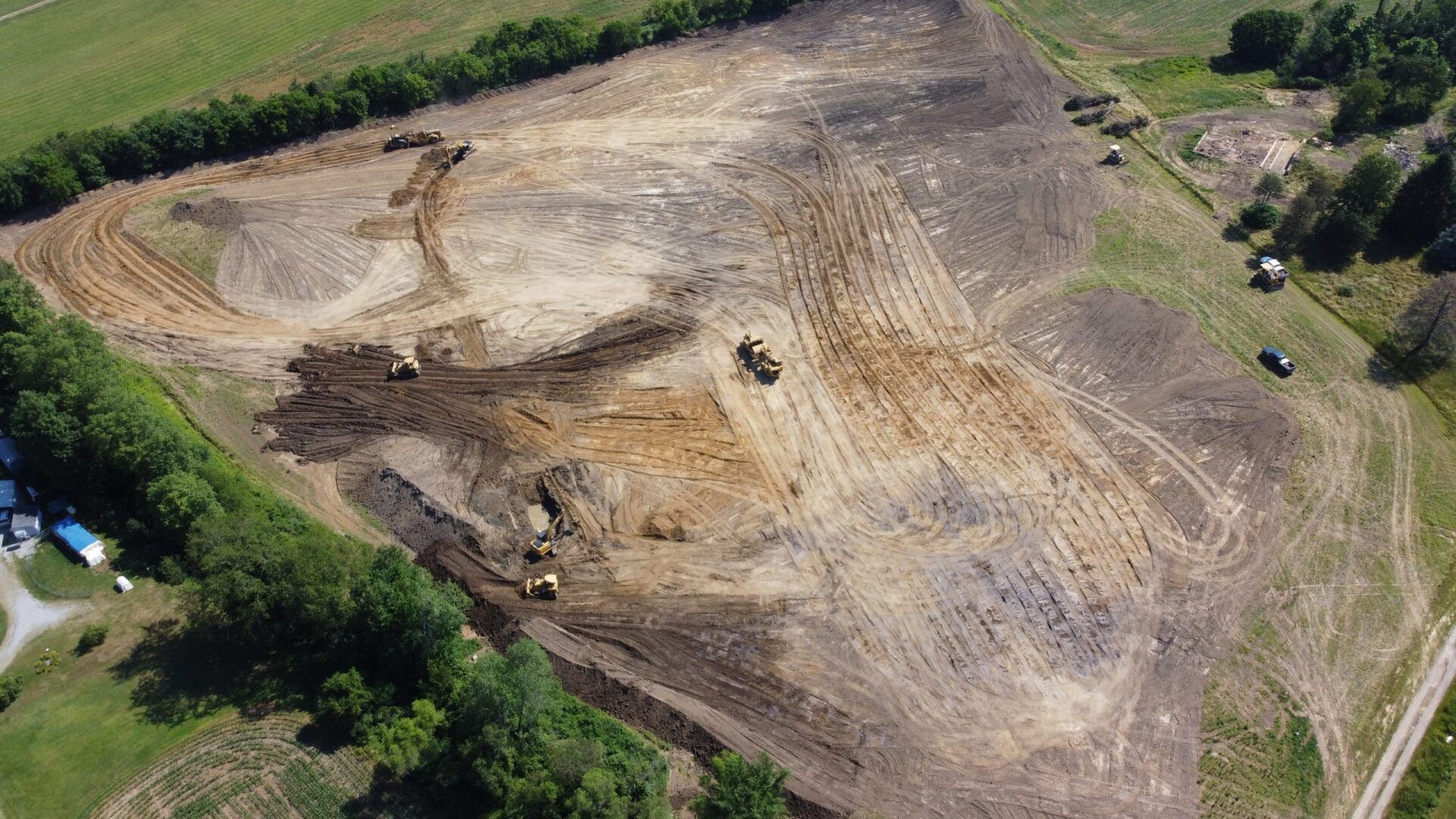 Aerial view of a deforested field with heavy machinery. Brown and black earth, green trees, and blue sky.
