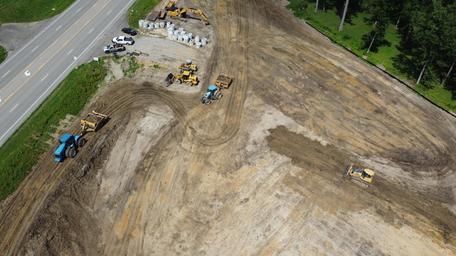 Construction site with multiple bulldozers working on a large plot of brown earth near a road and trees.