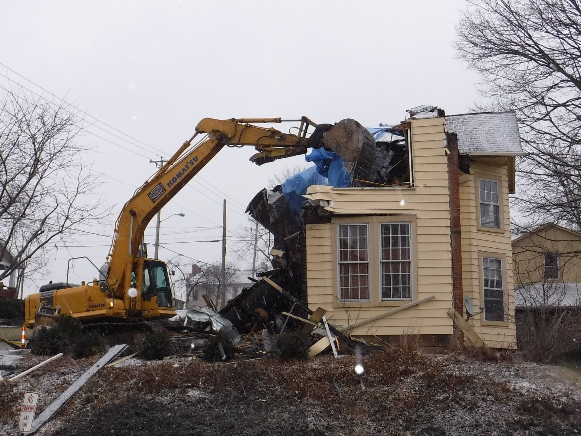 A yellow excavator demolishes a two-story house, snowy scene.