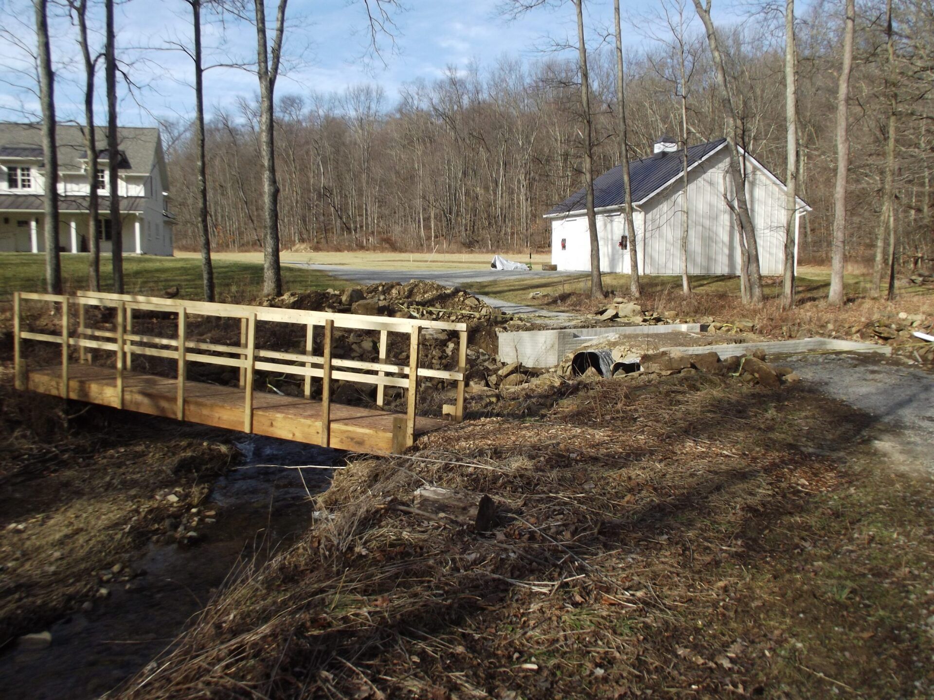 Wooden bridge over a creek in a wooded area, with a building and house visible.