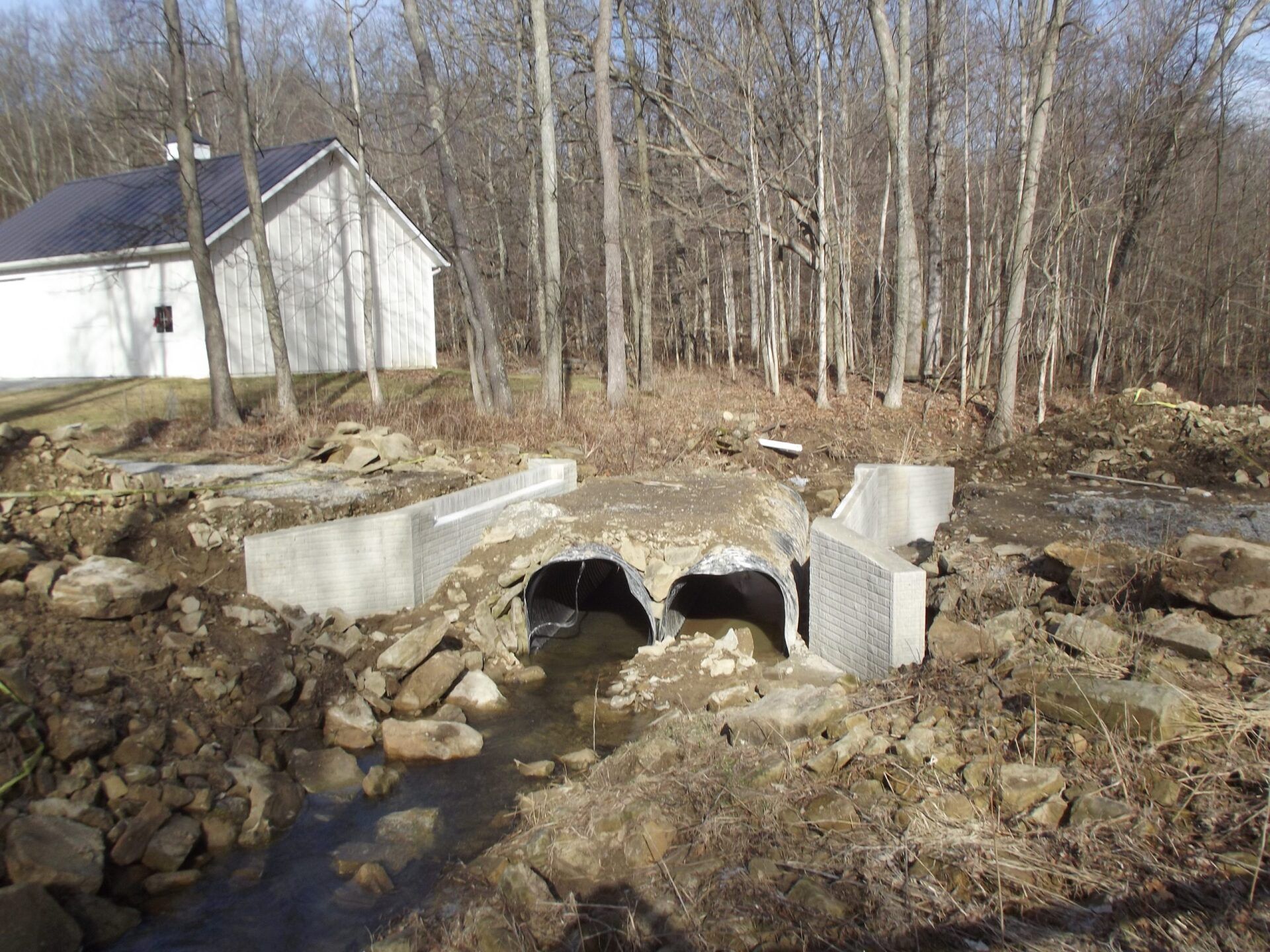 Concrete culvert over a stream with rocks and dirt, trees in the background, and a white building to the left.