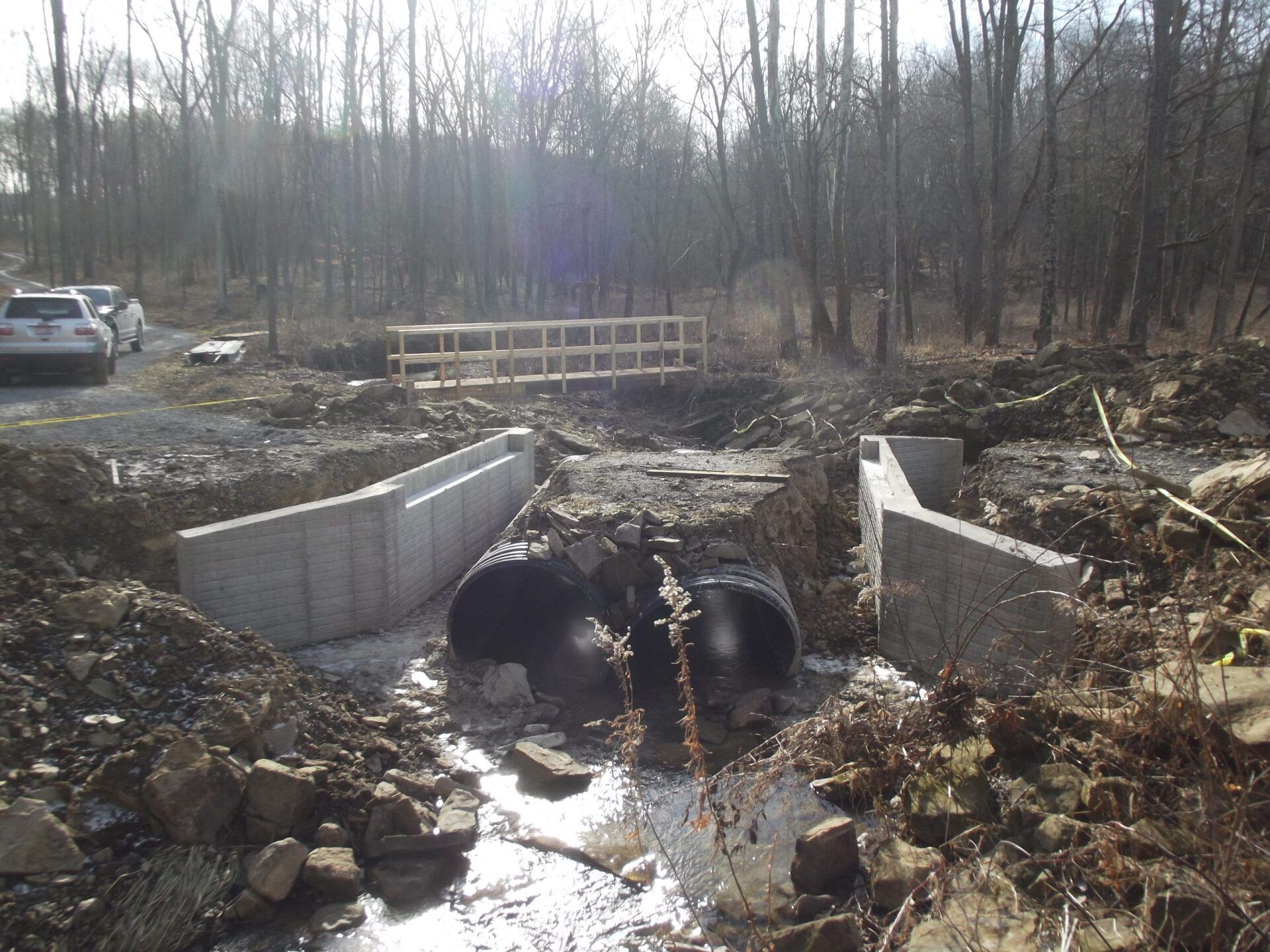 Construction site with concrete structures, culvert, and wooden bridge in a wooded area.