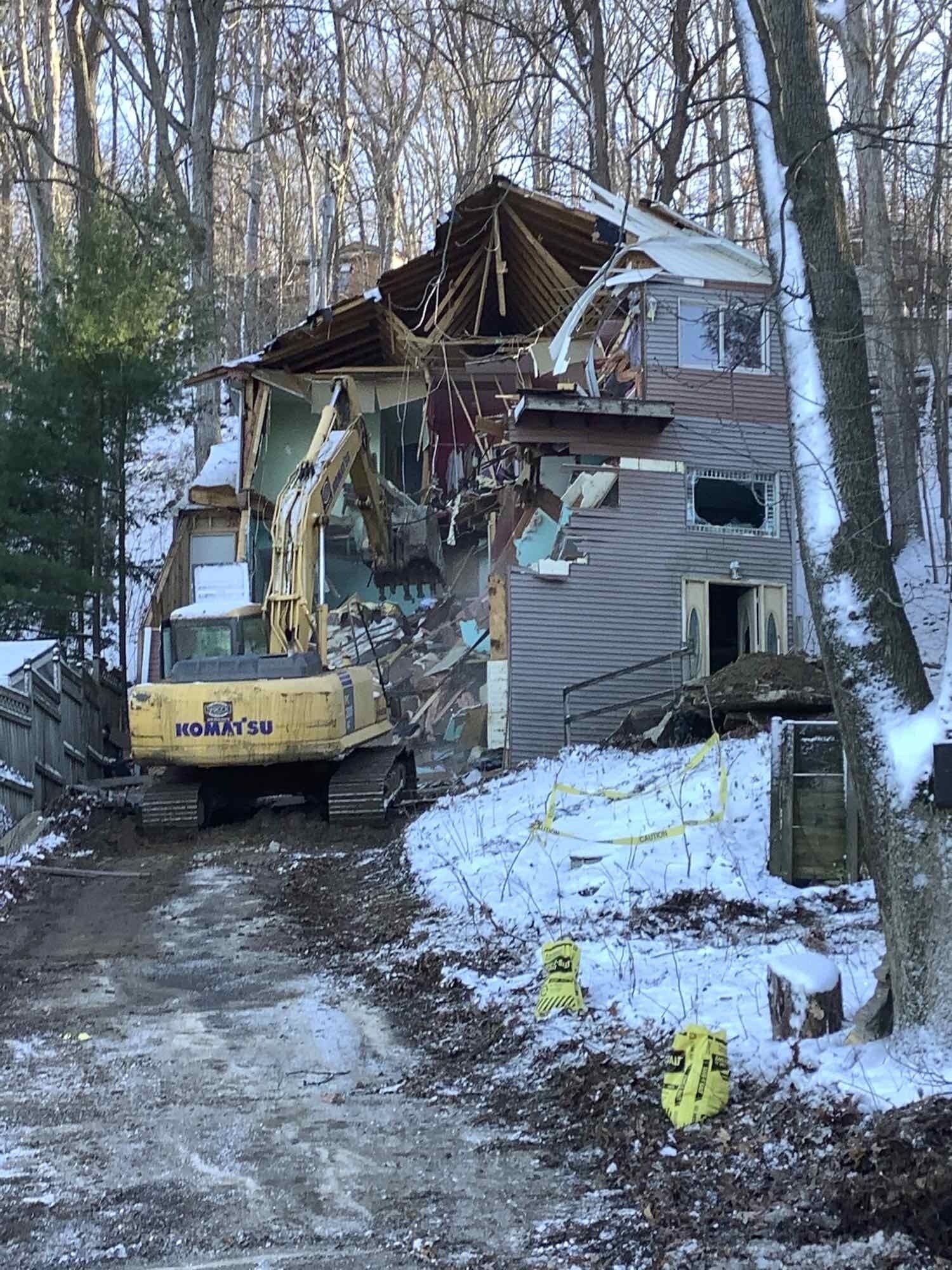 An excavator demolishes a house in a snowy, wooded area. Yellow caution tape and debris are visible.