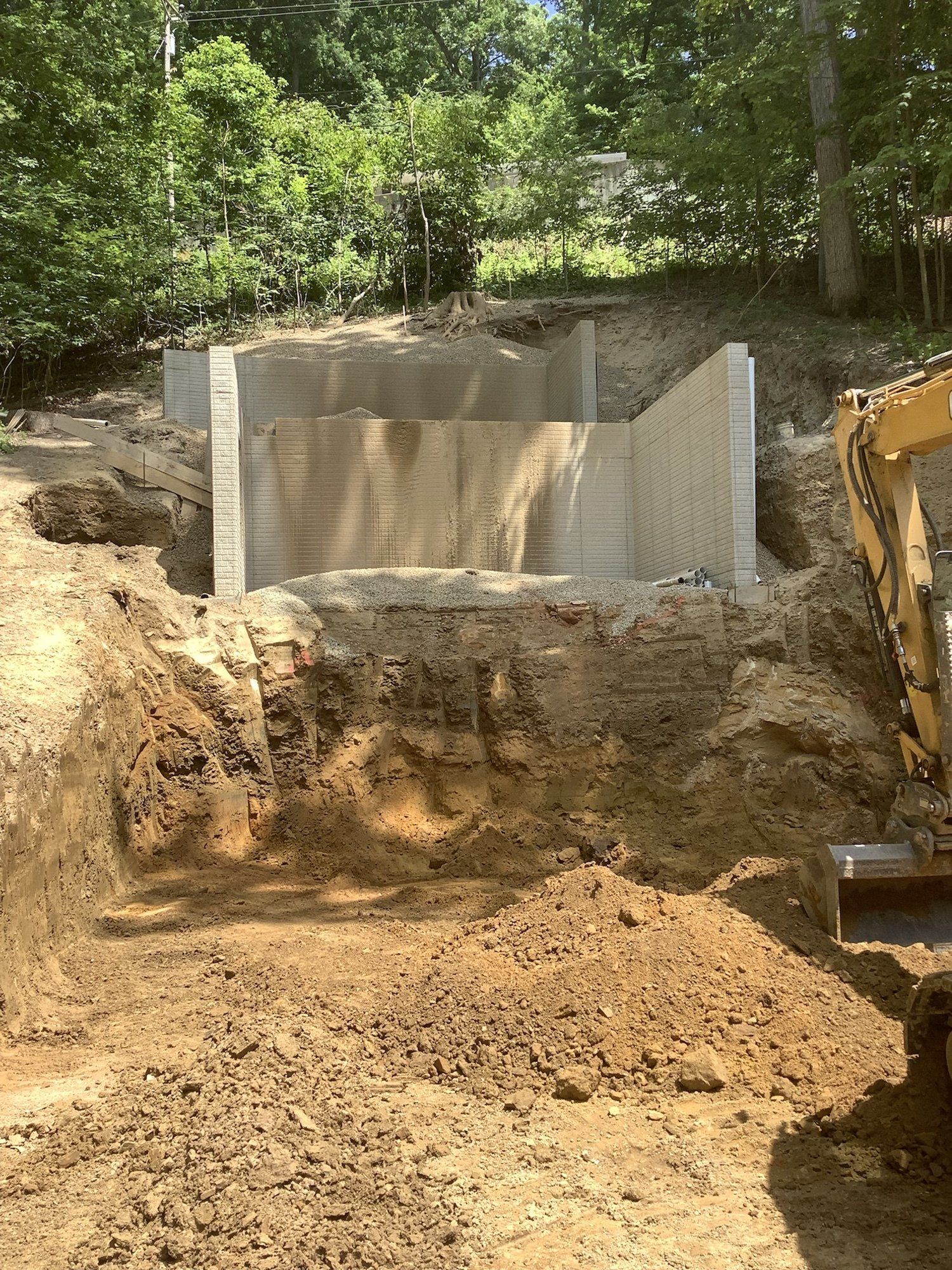 Excavation site with concrete retaining walls; construction in progress. Soil surrounds walls, excavator on right.