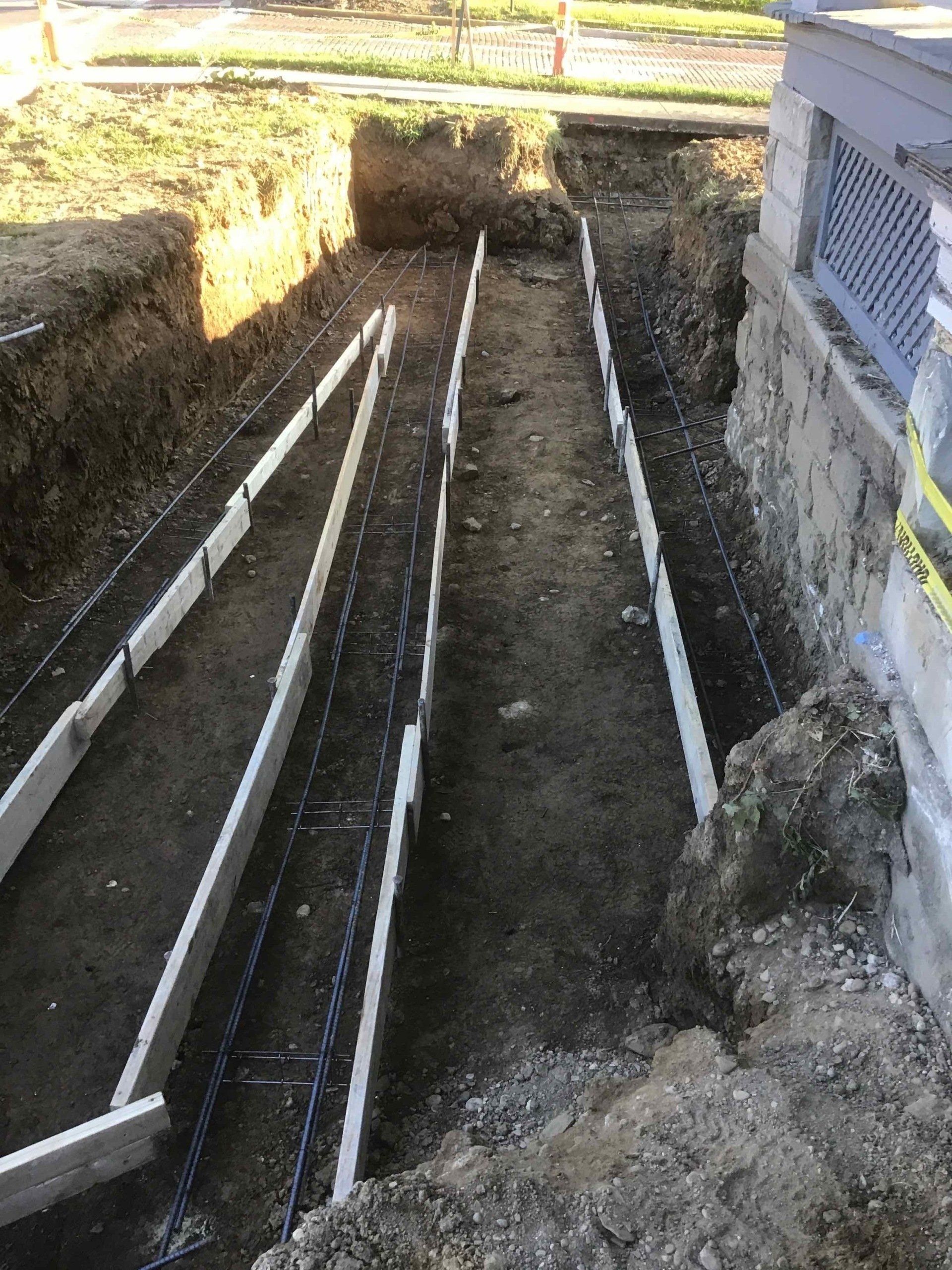 Construction site: trench with concrete forms and rebar along a building foundation.
