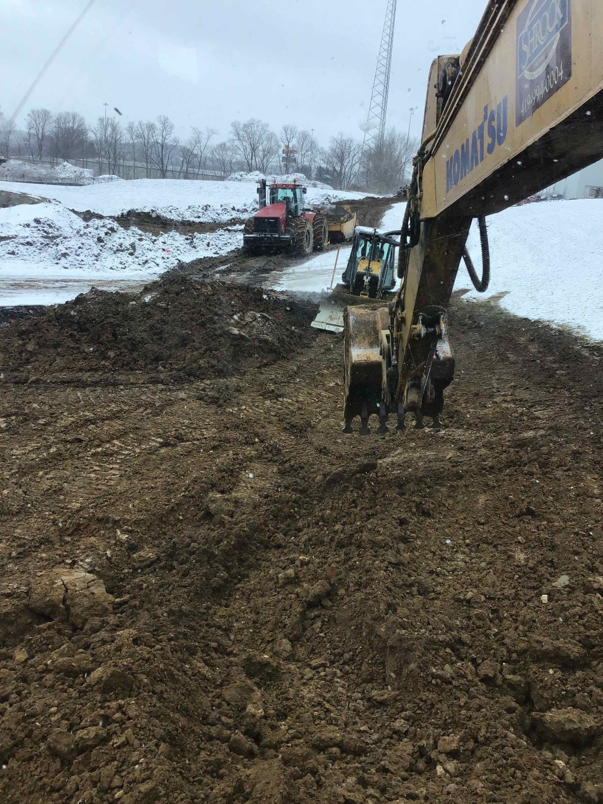 An excavator digs in muddy ground, with tractors and snowy ground in the background.