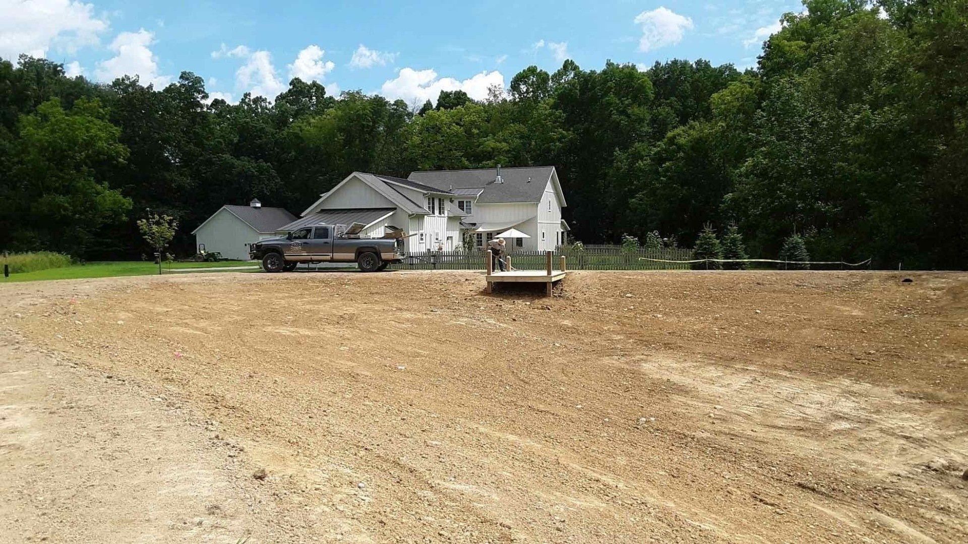 A house and trees sit behind a large mound of dirt with a truck and porta-potty in the middle, and blue sky.